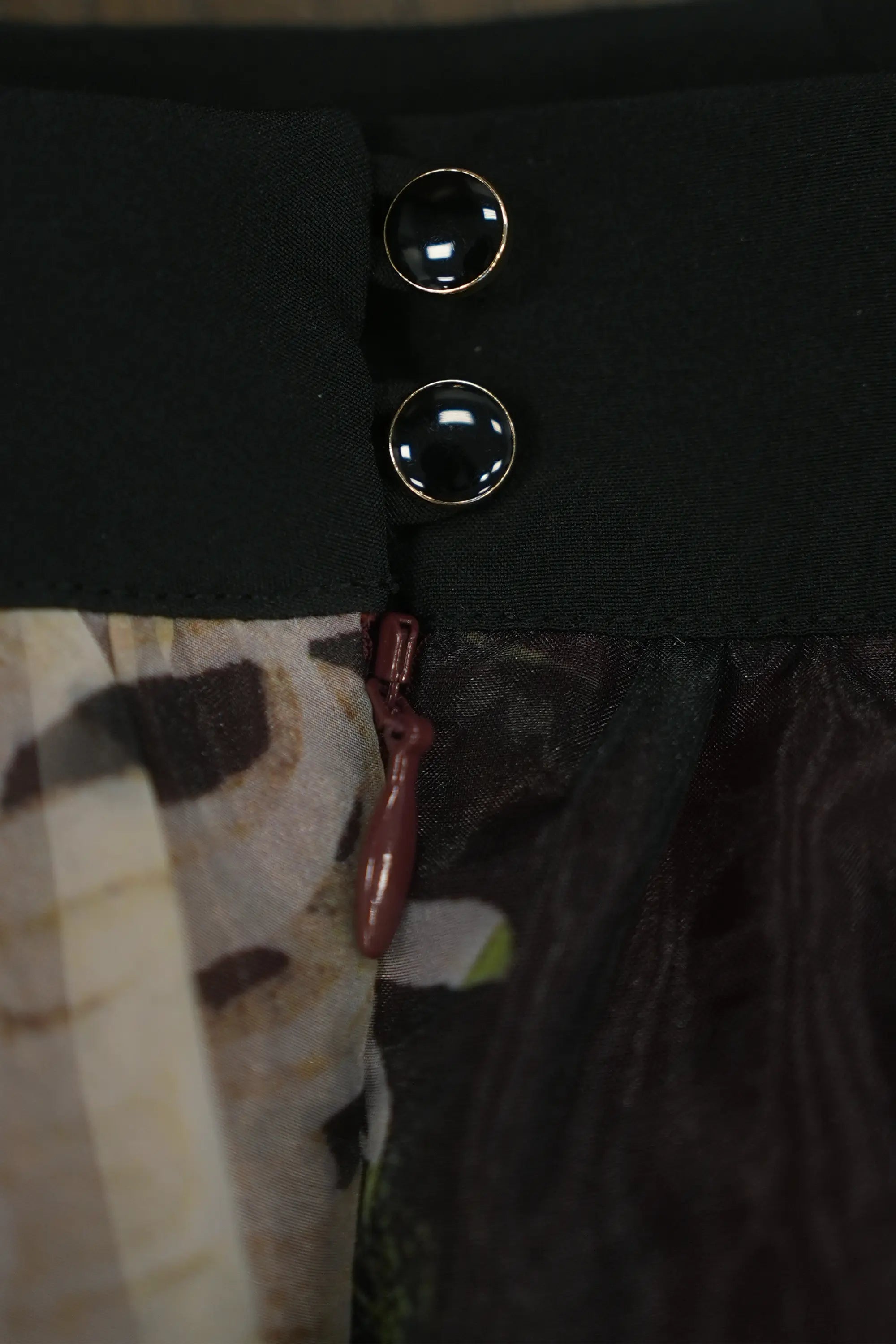 Close-up of skirt waistband featuring glossy button details and concealed side zip, highlighting precise construction, secure closure, and the contrast between structured waistband and sheer floral overlay