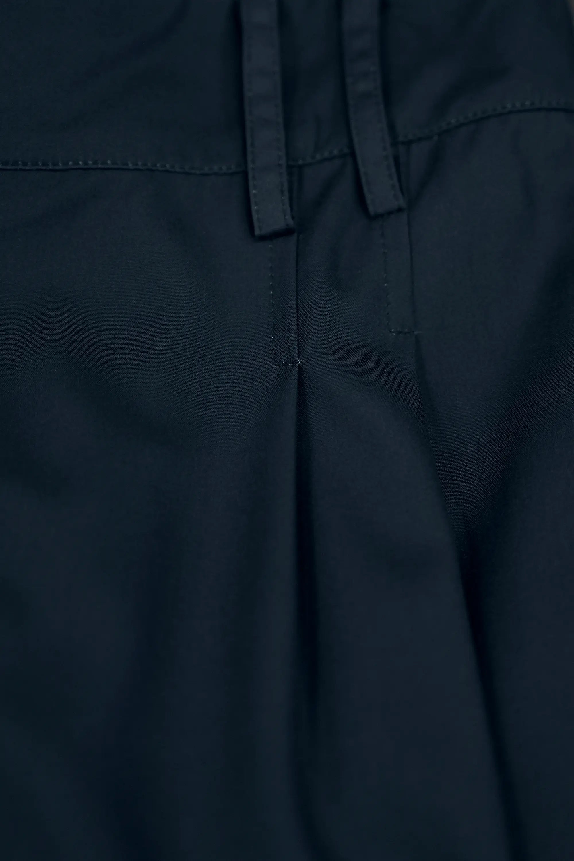 Close-up of dark pleated slit pants highlighting belt loops, structured waistband seam, and precise front pleat detailing, showcasing clean construction and tailored finishing