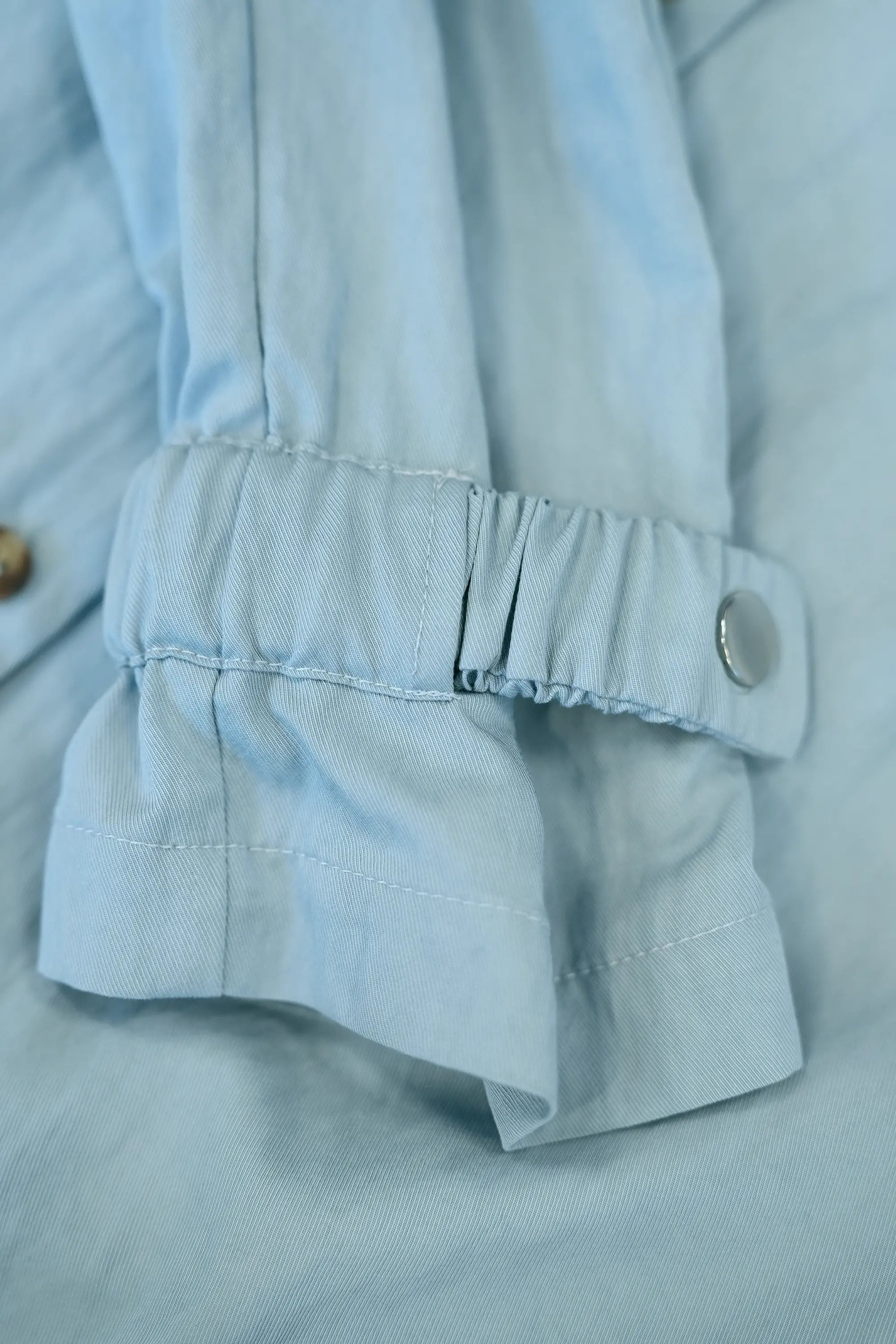Close-up of light blue shirt dress sleeve featuring adjustable elastic ruched cuff with button tab, highlighting gathered detailing, flexible fit, and clean stitching finish
