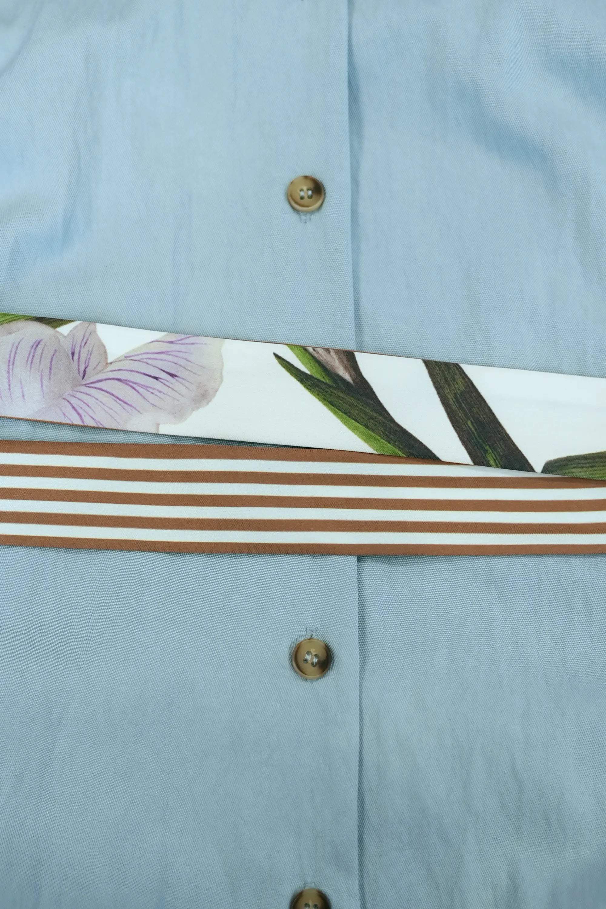 Close-up of light blue shirt dress waist detail featuring contrast striped sash belt with printed floral panel, highlighting layered textures and button down front