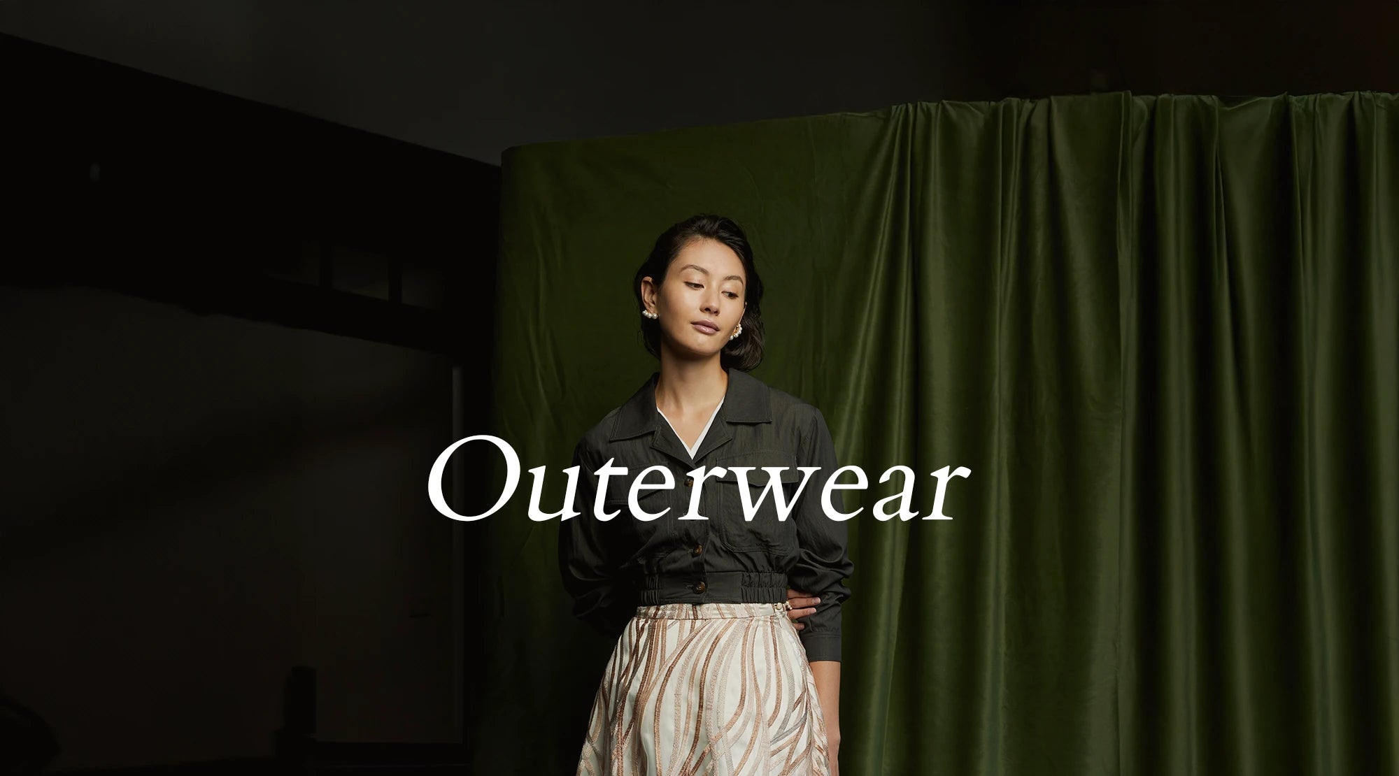 Outerwear collection banner, desktop version, with a woman in a dark utility jacket and striped skirt standing against a green curtain