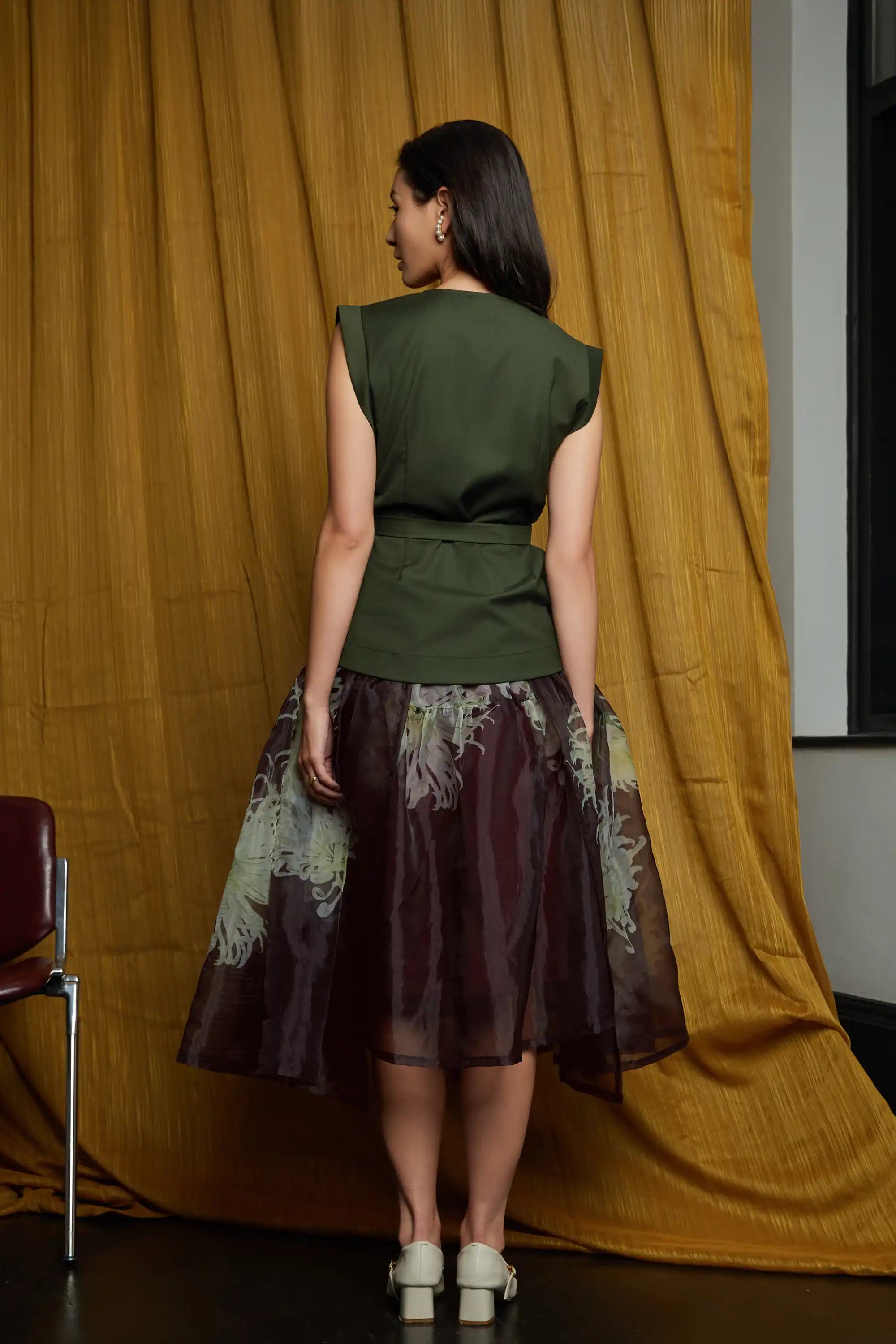 Full back view of model wearing deep green structured top with a clean fitted back and waist sash tie, paired with a sheer floral overlay skirt, showcasing a defined waistband, layered transparency, and a softly flowing silhouette