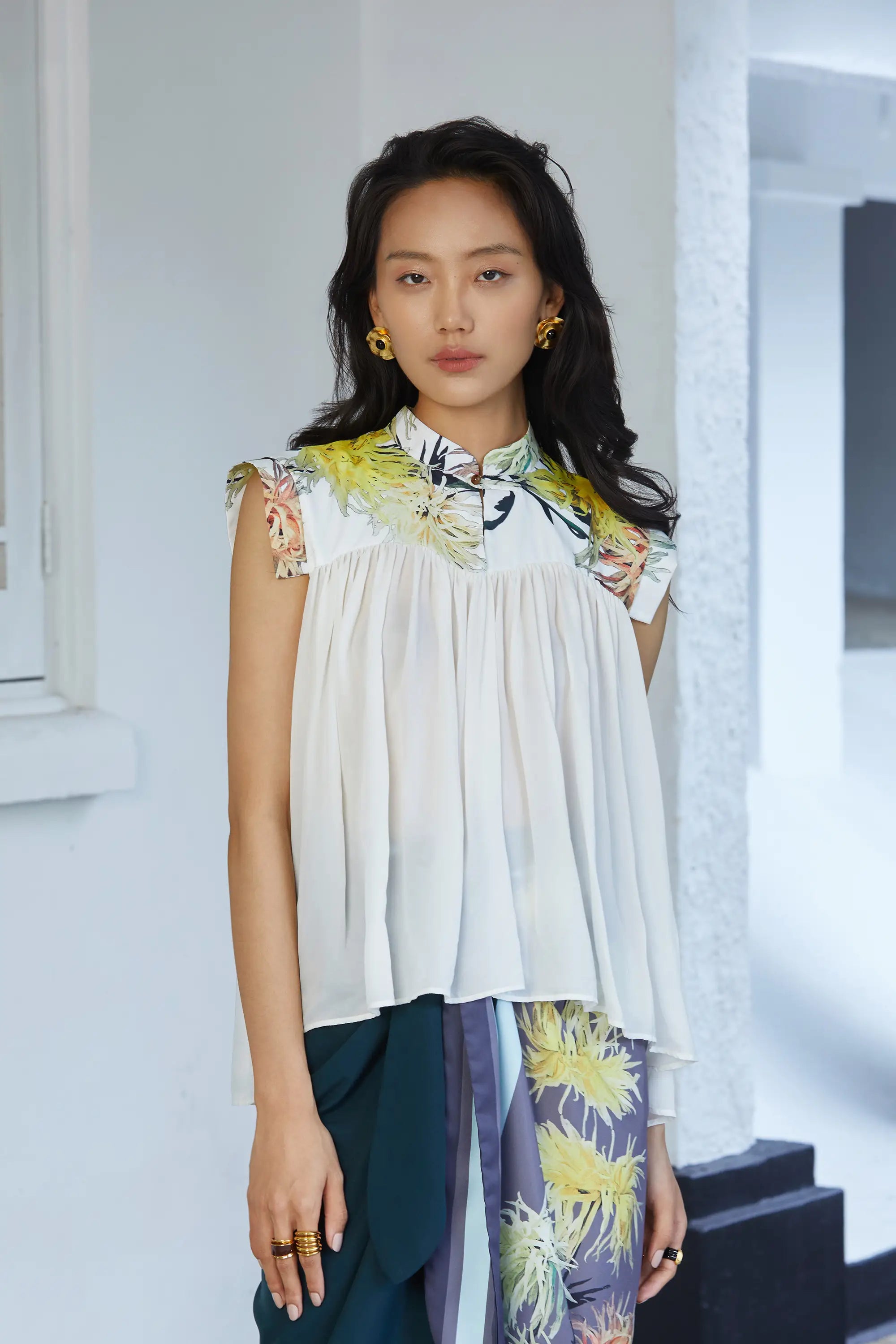 Sleeveless white blouse with a softly gathered, airy silhouette, featuring a high printed collar with floral motifs, extended shoulder caps, and a fluid drape over the torso.