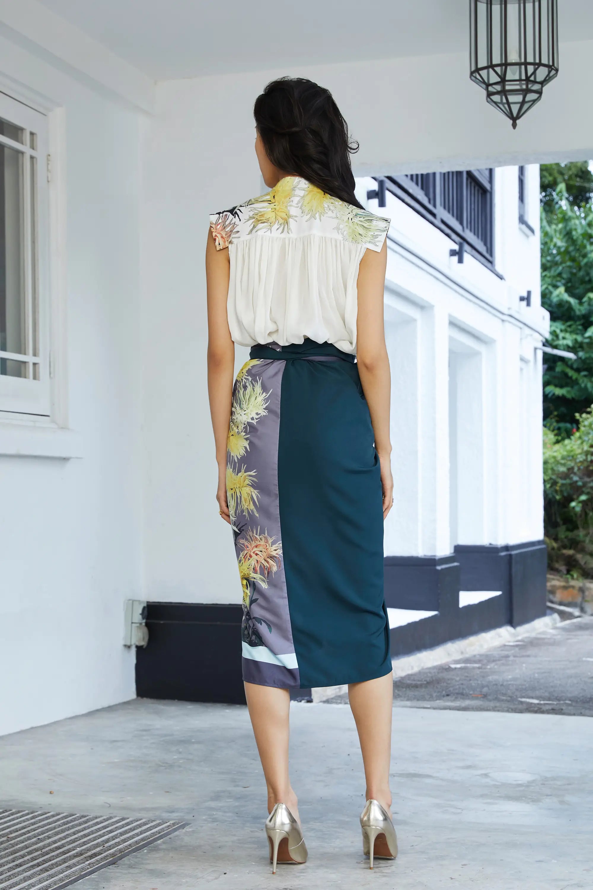Back view of white sleeveless blouse with gathered drape and floral printed high collar, paired with a midi wrap skirt featuring contrast panels in deep green and chrysanthemum print, finished with an adjustable waist tie.