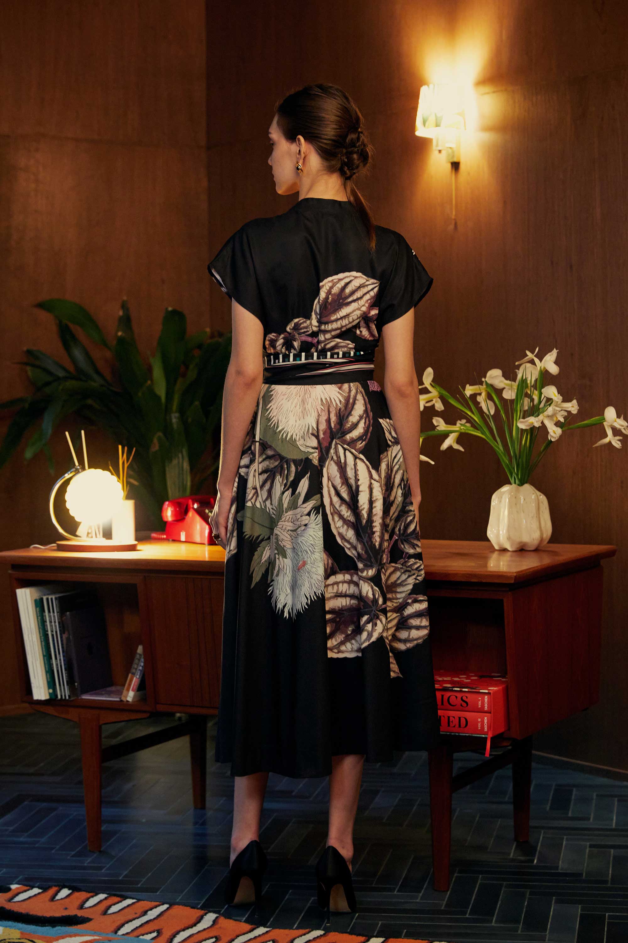 Back view of model wearing the BEHOLD Blended Collar Wrap Midi Dress in Stokesia print, standing in a warm wood-panelled room with mid-century furnishings and fresh florals.