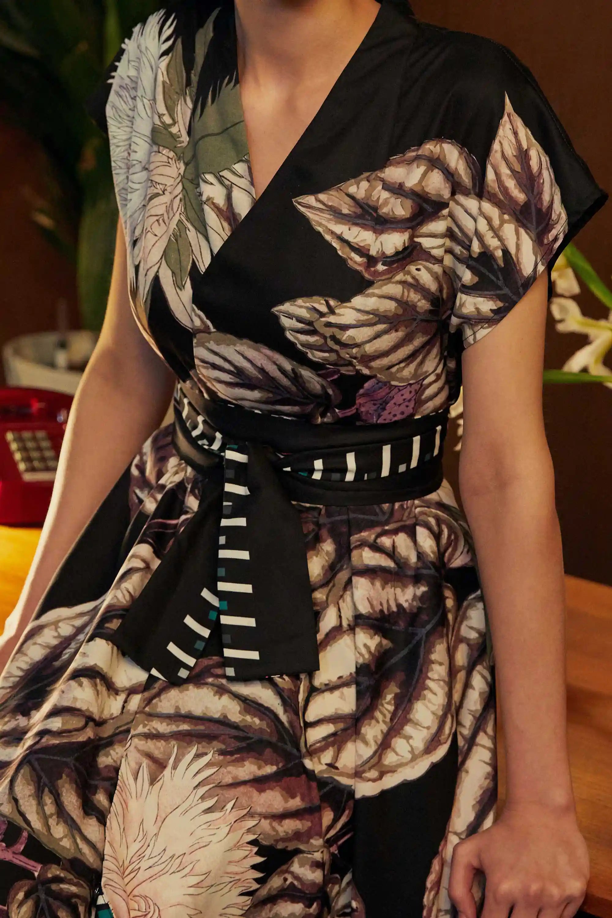 Close-up of model in the BEHOLD Blended Collar Wrap Midi Dress with Stokesia print, showcasing the pleated waist, obi-style belt, and painterly florals in ivory and plum. Captured in a vintage interior with a red rotary phone.