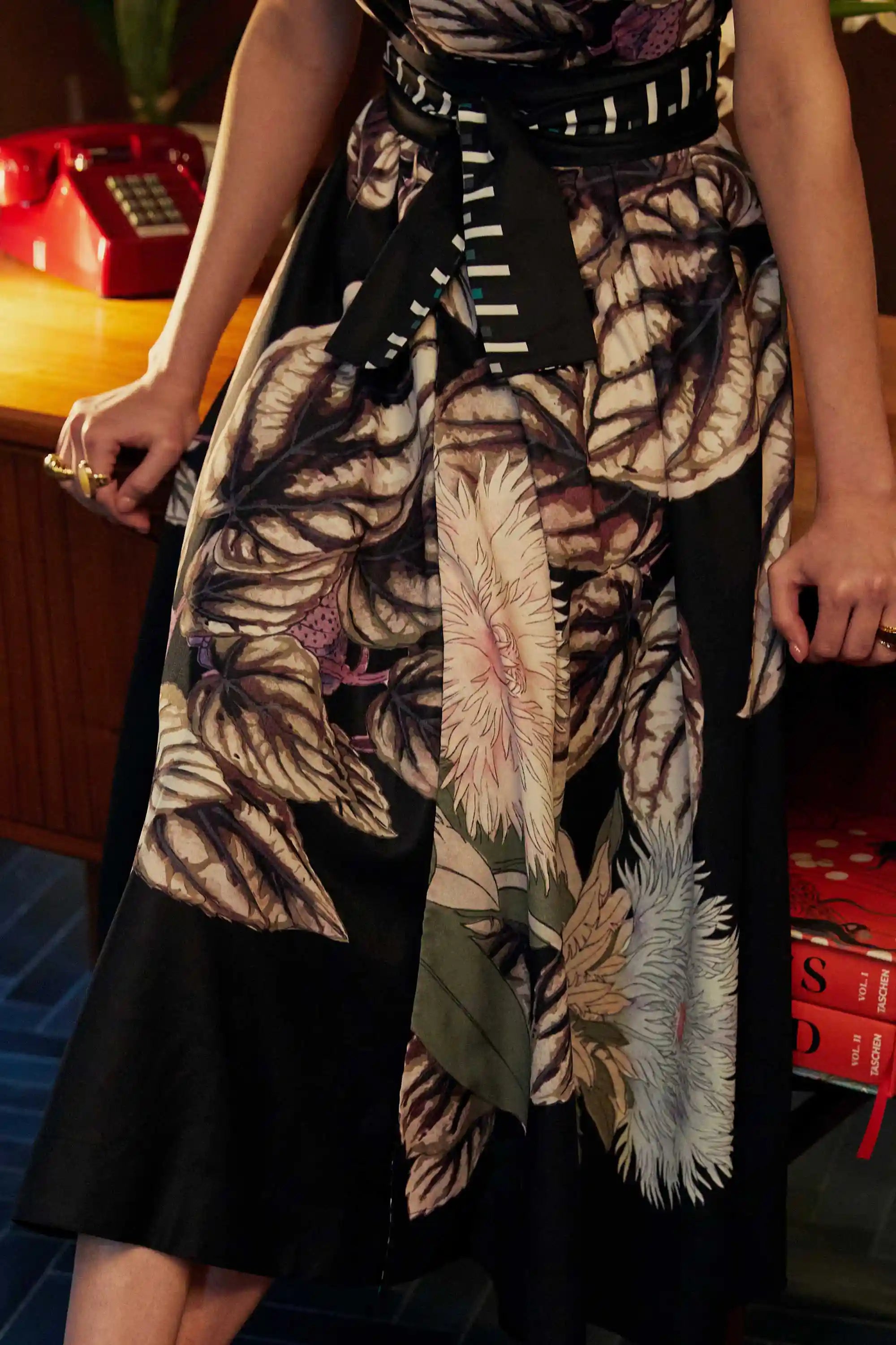 Close-up of the BEHOLD Blended Collar Wrap Midi Dress in Stokesia print, highlighting painterly florals, structured pleats, and a graphic obi belt. Captured indoors with a red rotary phone and vintage books in the background.