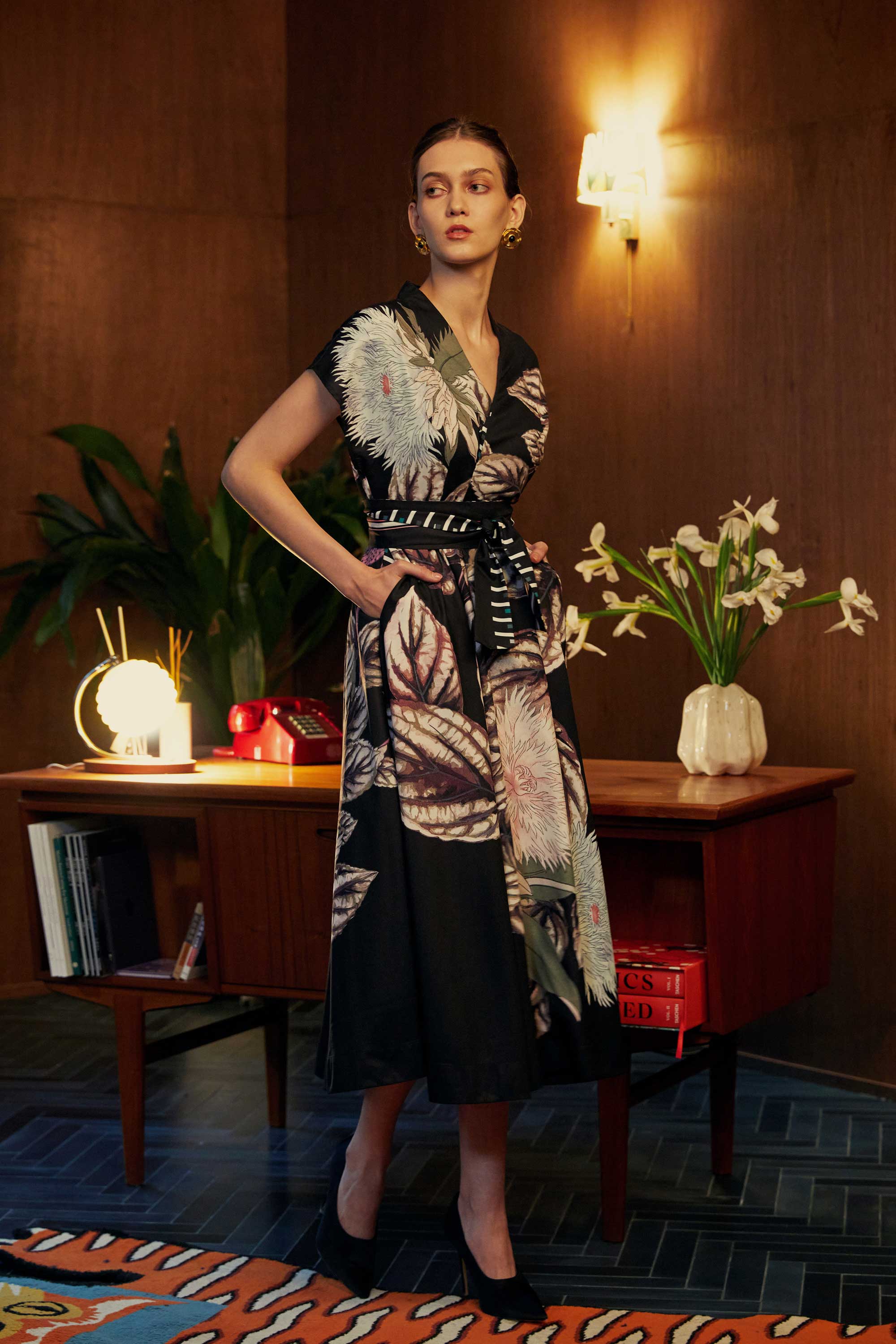 Model in the BEHOLD Blended Collar Wrap Midi Dress in Stokesia print, standing with hands in pockets in a warmly lit wood-panelled room with lush plants and mid-century décor.