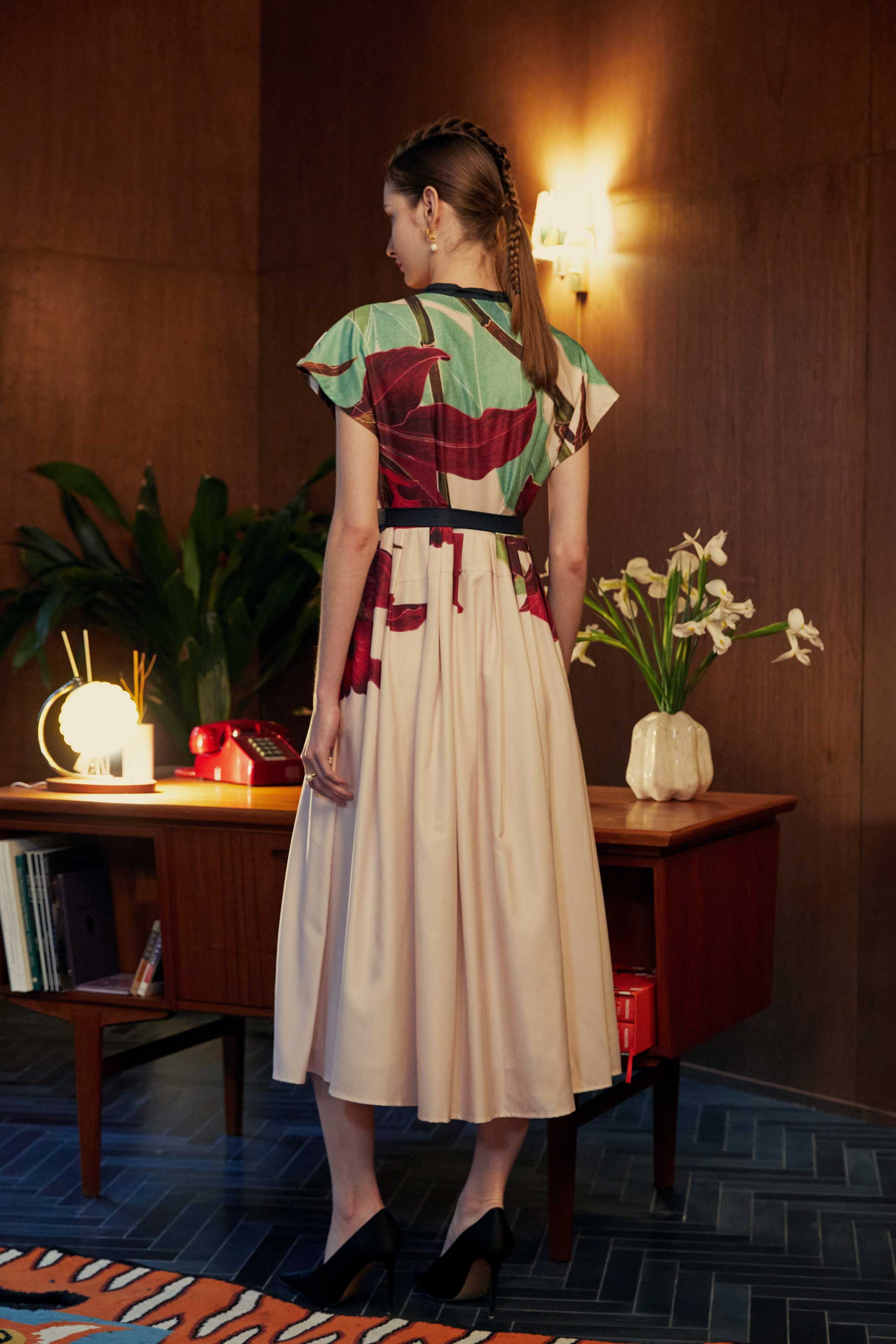 Back view of the BEHOLD Chinese Collar Fit & Flare Maxi Dress in Laelia print, featuring oversized orchid motifs in emerald and crimson. Shot in a wood-panelled room styled with vintage décor, floral accents, and warm ambient lighting.