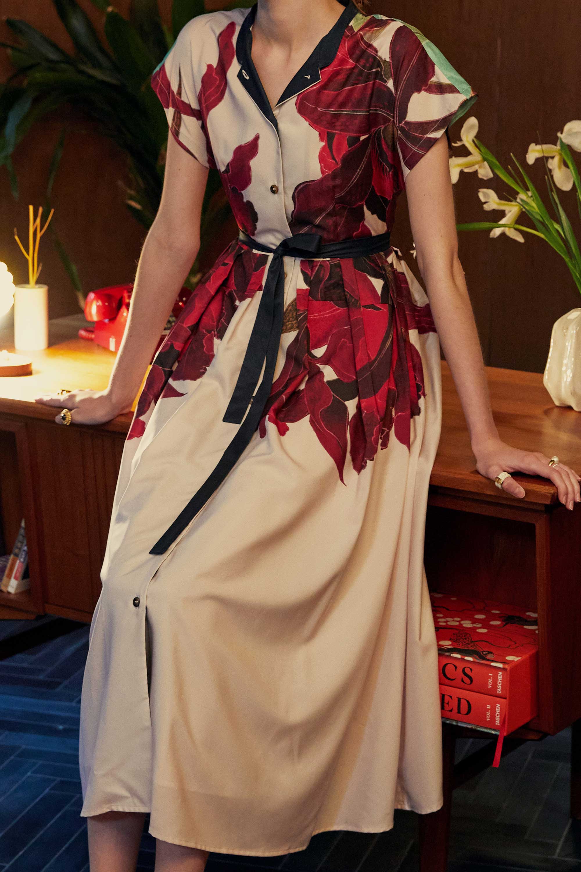Close-up of the BEHOLD Chinese Collar Fit & Flare Maxi Dress in Laelia print, highlighting the oversized crimson orchid motifs against a cream base, with a black sash at the waist. Shot indoors with wood-panelled furniture, a tiger-striped rug, and ambient lighting
