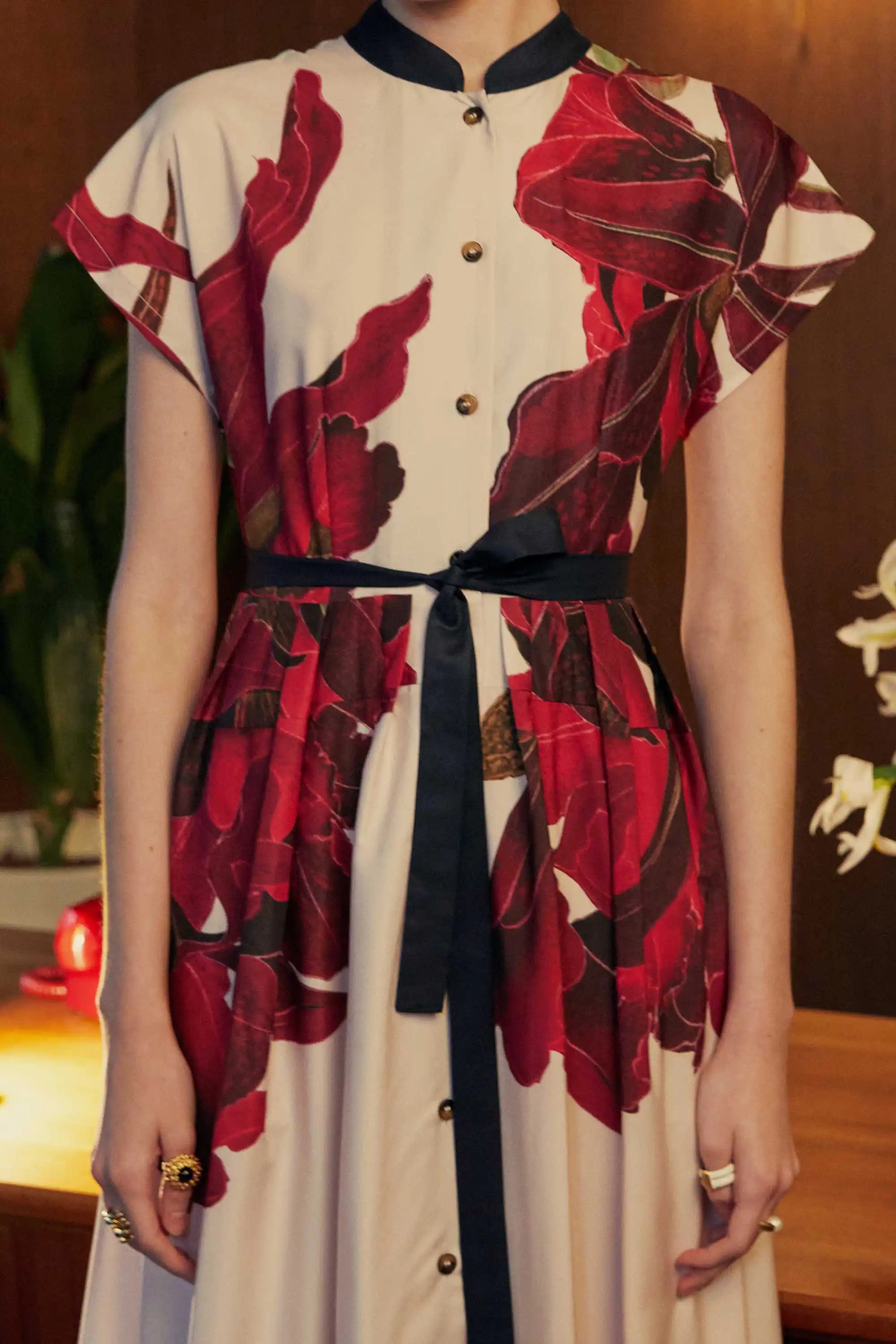Cropped close-up of the BEHOLD Chinese Collar Fit & Flare Maxi Dress in Laelia print, showcasing the mandarin collar, tortoise-shell buttons, and bold crimson orchid motifs contrasted against a cream base. Styled with a black waist tie, captured in a warmly lit indoor setting.