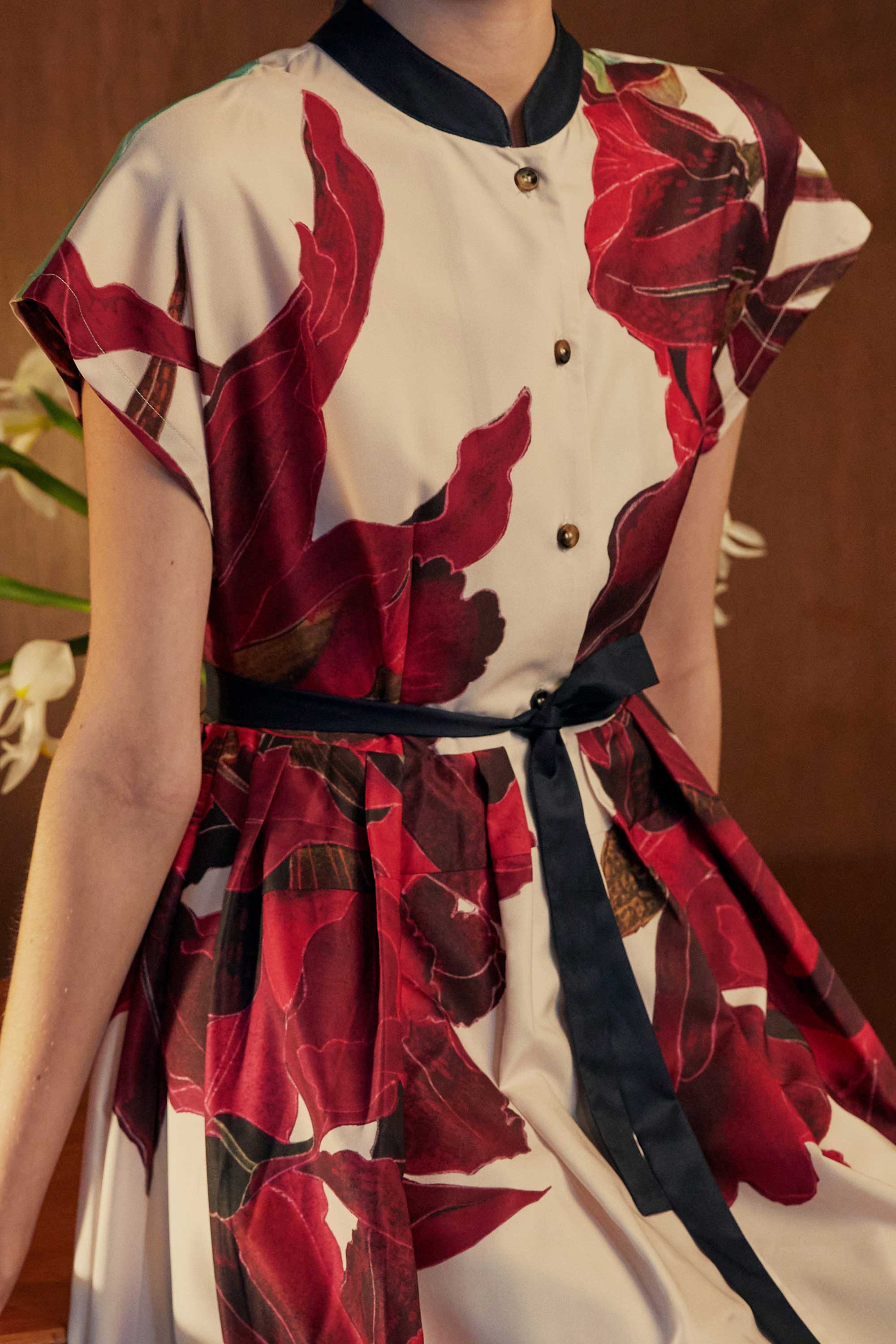 Close-up of the BEHOLD Chinese Collar Fit & Flare Maxi Dress in Laelia print, showcasing the sculptural crimson orchid motifs and contrast black mandarin collar. Pleated skirt and waist tie details are framed by warm ambient lighting and floral accents in the background.