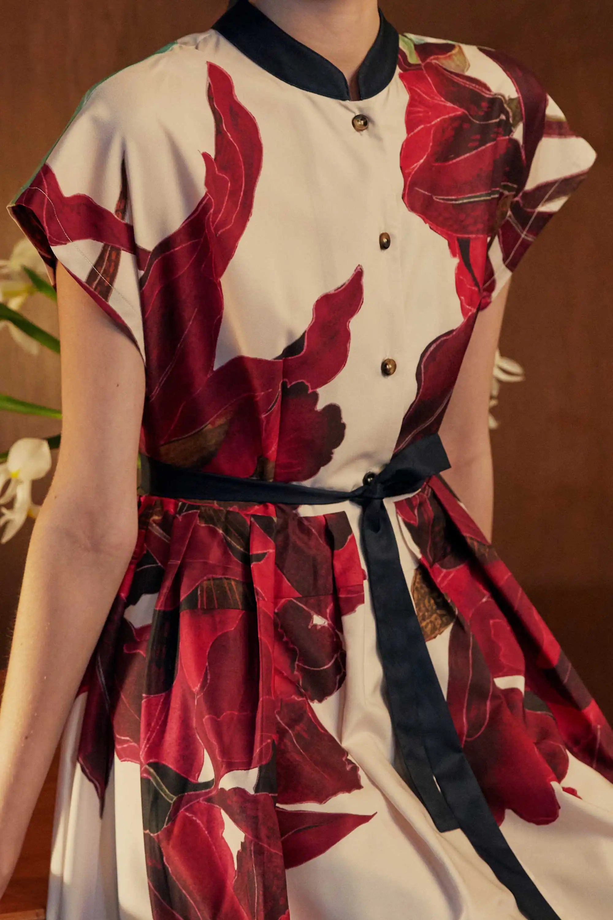 Close-up of the BEHOLD Chinese Collar Fit & Flare Maxi Dress in Laelia print, showcasing the sculptural crimson orchid motifs and contrast black mandarin collar. Pleated skirt and waist tie details are framed by warm ambient lighting and floral accents in the background.