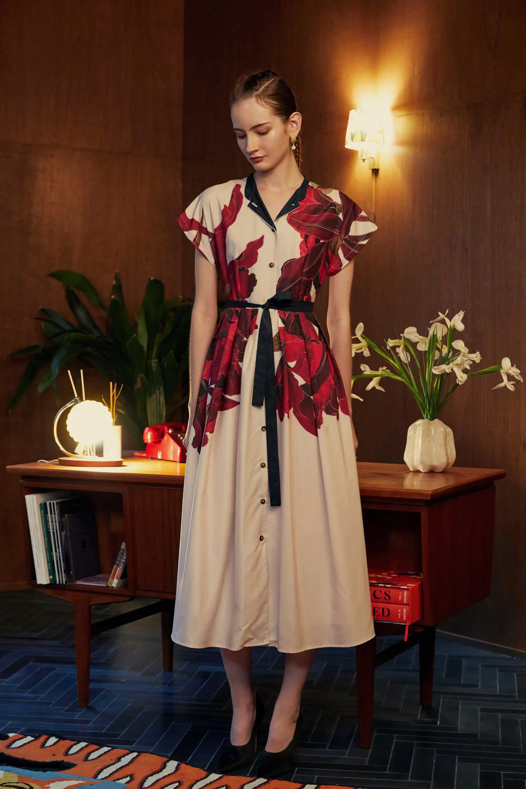 Model wearing the BEHOLD Chinese-Collar Fit-and-Flare Maxi Dress in Laelia orchid print—cream button-down maxi with black mandarin collar, flutter sleeves, and contrast tie-belt, styled indoors with wood-paneled backdrop.