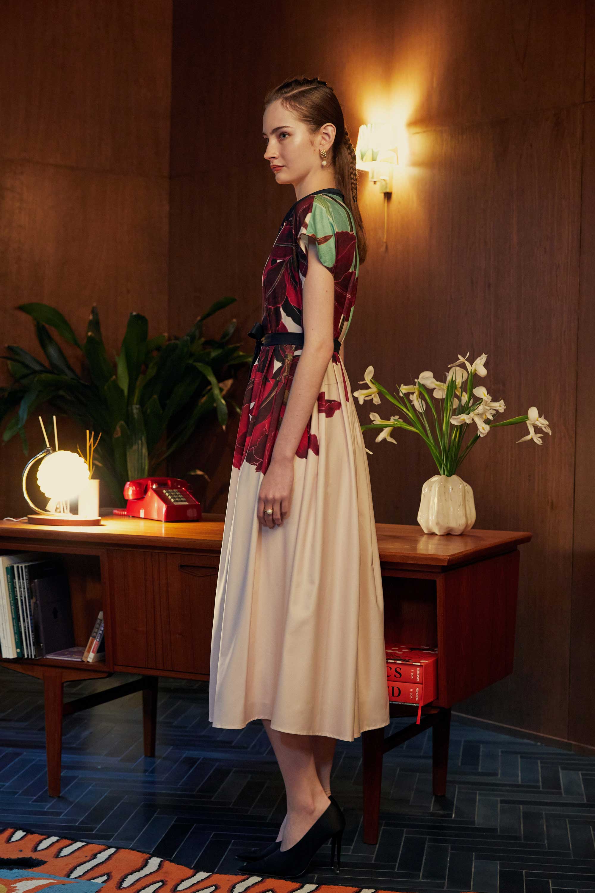 Side profile of model wearing the BEHOLD Chinese Collar Fit & Flare Maxi Dress in Laelia print, showcasing structured cap sleeves and an elegant flare silhouette. Set in a warm wood-panelled room with floral decor and vintage-inspired furnishings.