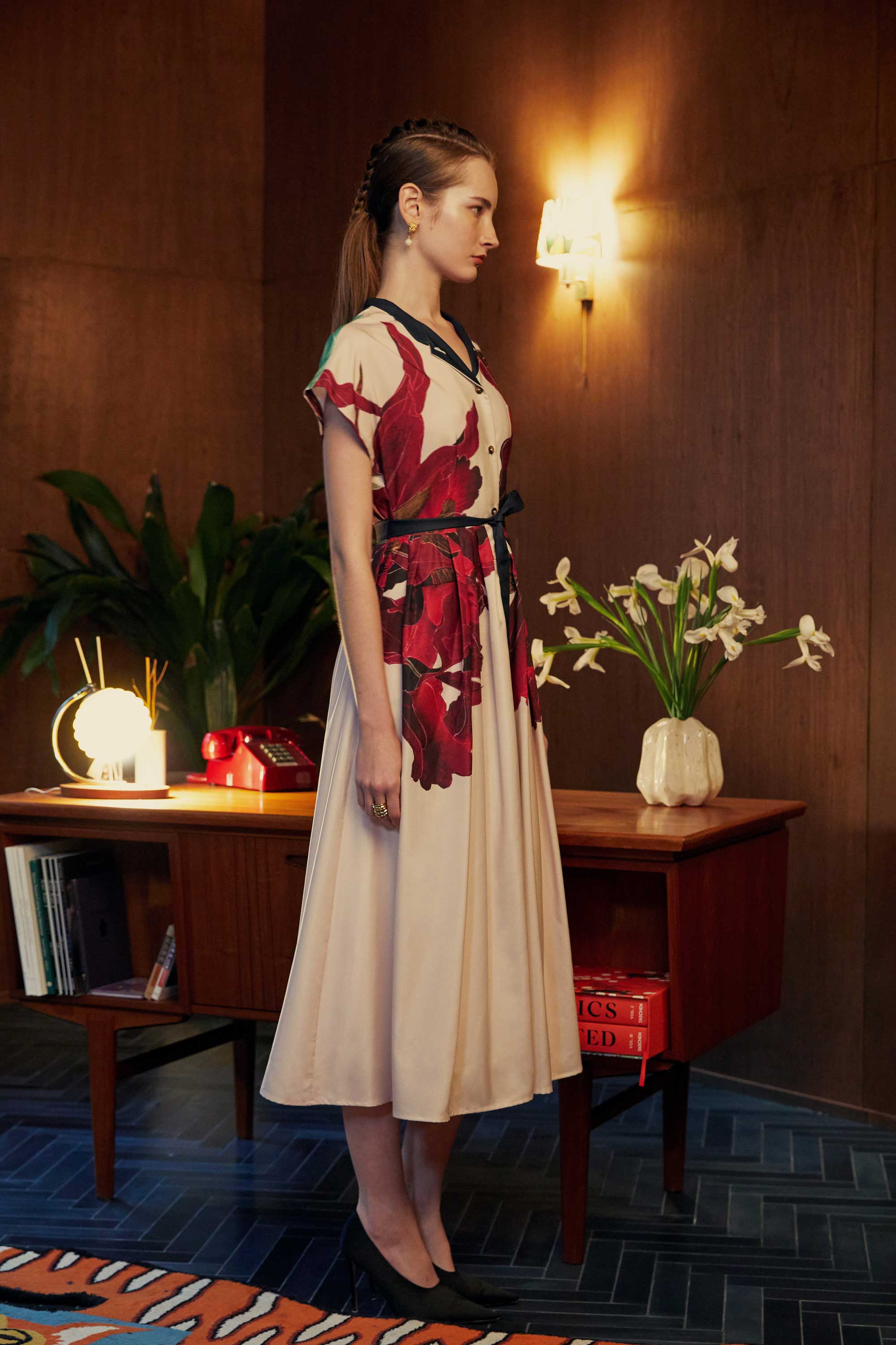 Side view of model in the BEHOLD Chinese Collar Fit & Flare Maxi Dress in Laelia print, featuring a structured silhouette with cap sleeves and bold orchid motifs. Photographed in a warm-toned wood-panelled room with floral accents and vintage décor.