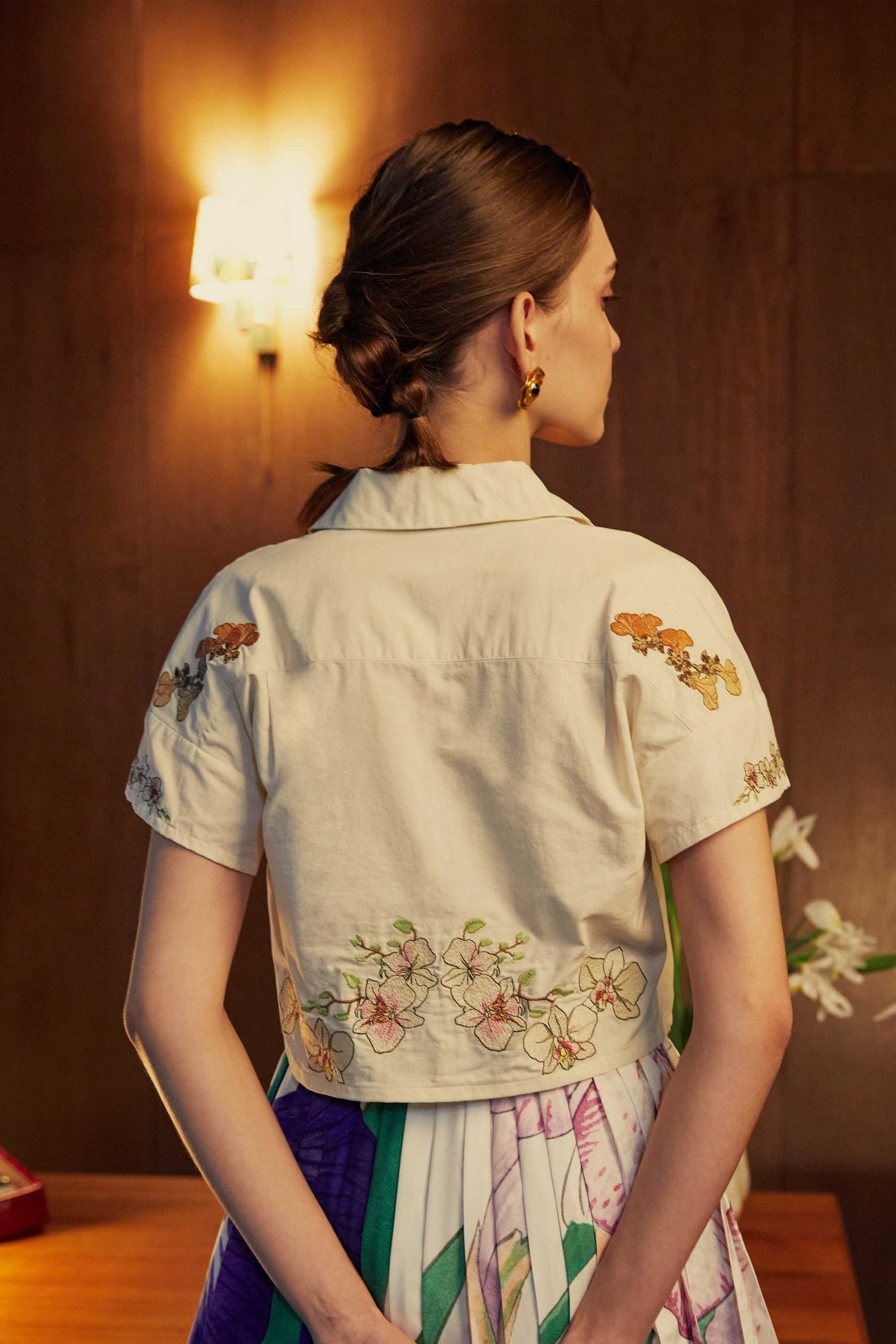 Back view of a cropped Cuban shirt with intricate floral embroidery, styled with pleated skorts in bold stripes, captured under warm lighting in a wood-panelled room.