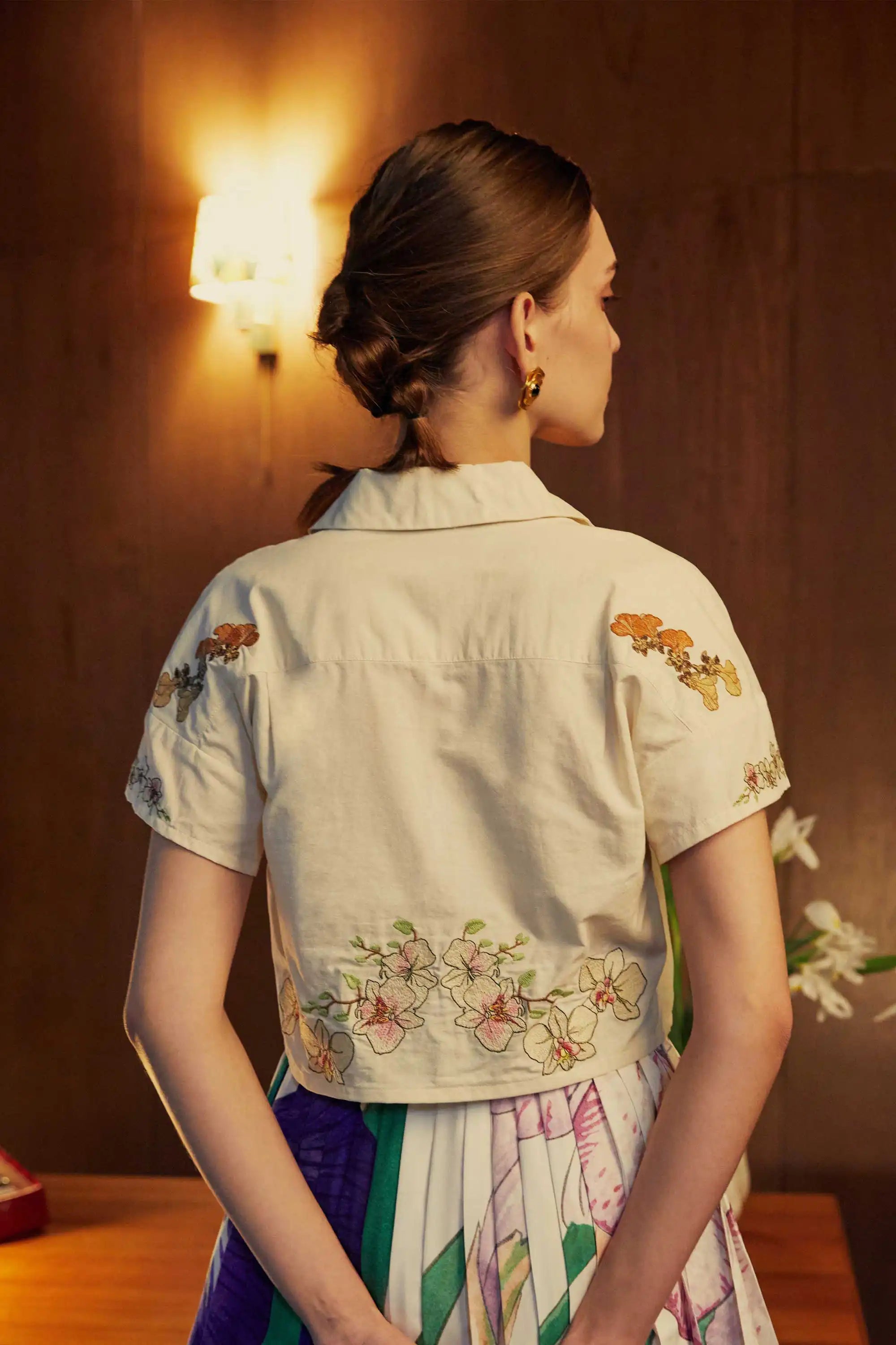 Back view of a cropped Cuban shirt with intricate floral embroidery, styled with pleated skorts in bold stripes, captured under warm lighting in a wood-panelled room.