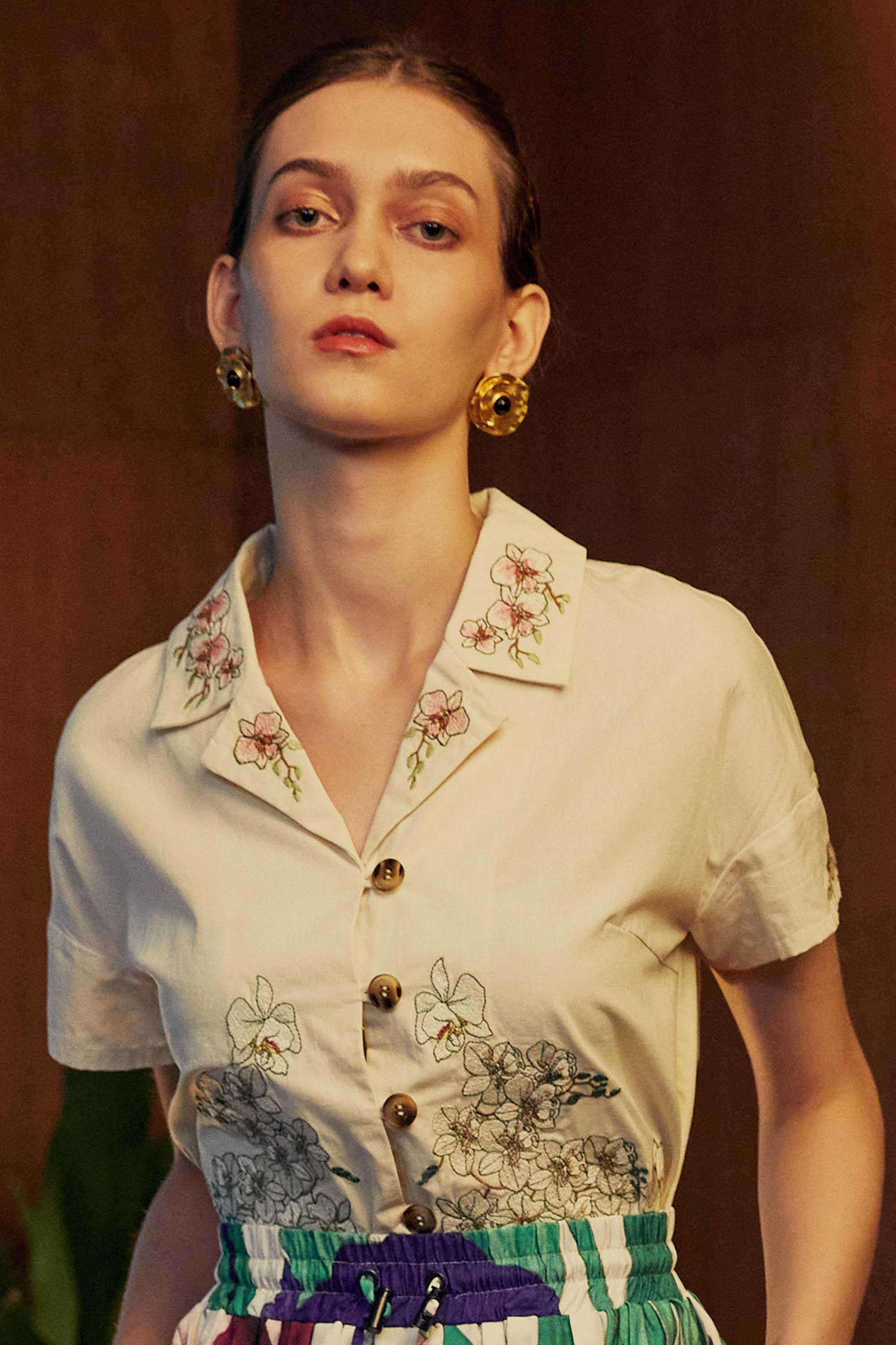 Close-up of a woman wearing a cropped Cuban shirt with floral embroidery and wooden buttons, styled with pleated Iris skorts in a wood-panelled interior.