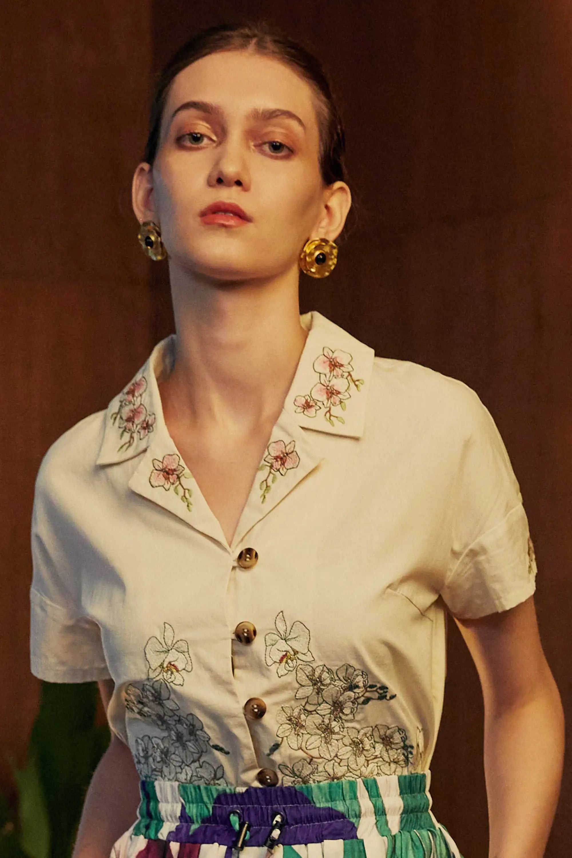Close-up of a woman wearing a cropped Cuban shirt with floral embroidery and wooden buttons, styled with pleated Iris skorts in a wood-panelled interior.
