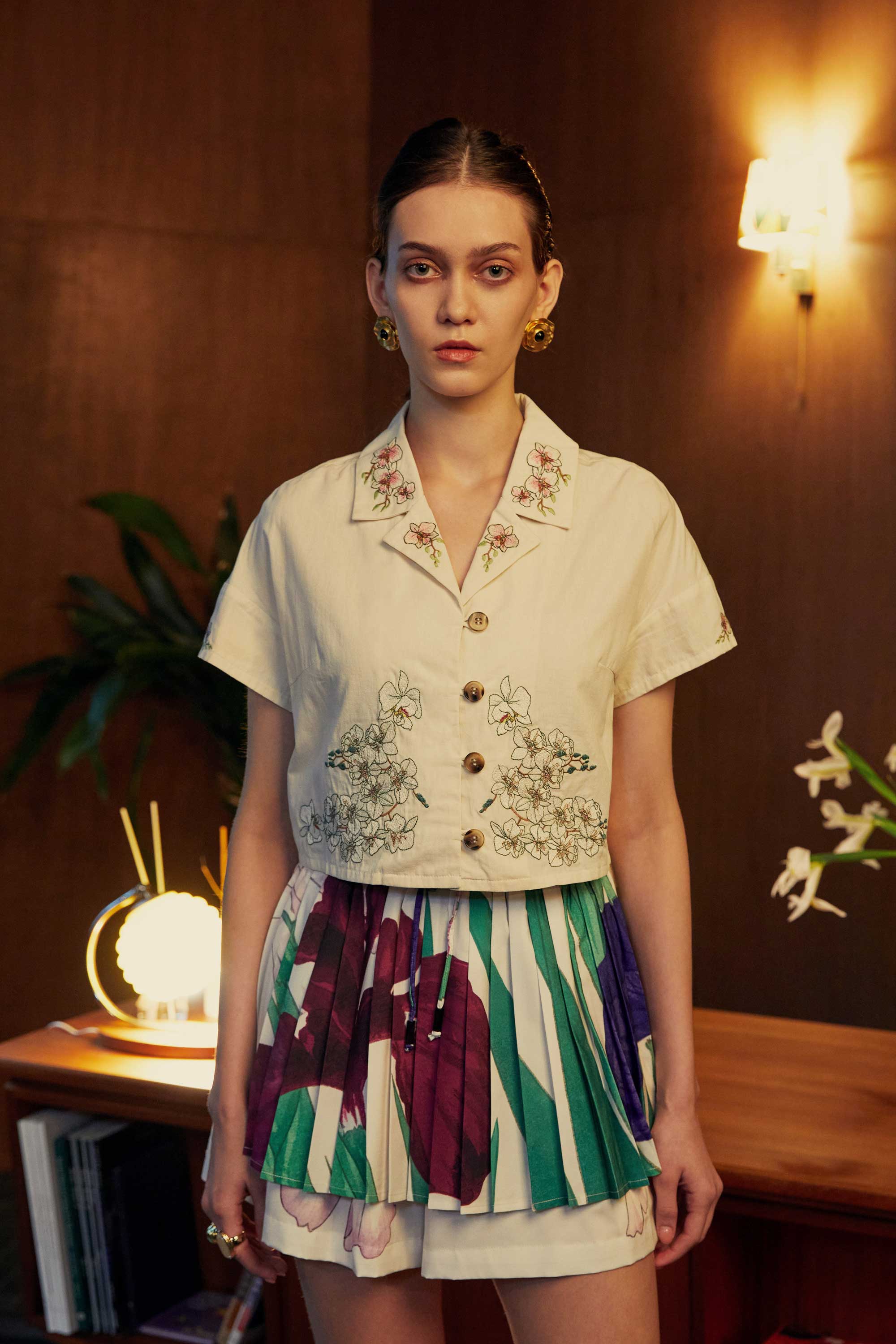 Model in a cropped Cuban shirt embroidered with delicate threadwork, paired with a pleated colour-blocked skorts in emerald, plum, and cream. Captured in a warm, mid-century styled room with soft lighting and tropical florals.