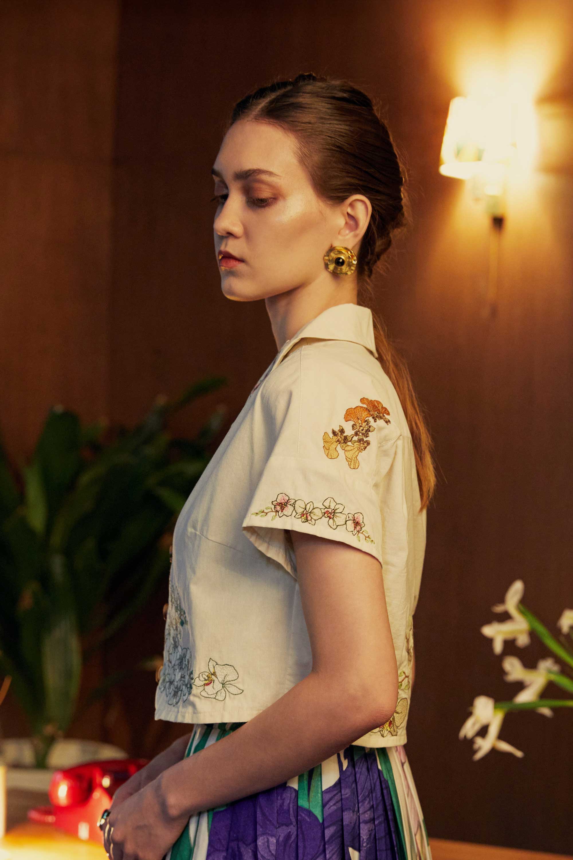 Side view of a model in a cropped Cuban shirt with floral embroidery, paired with pleated iris-print skorts — photographed indoors with golden-hour lighting and mid-century décor.