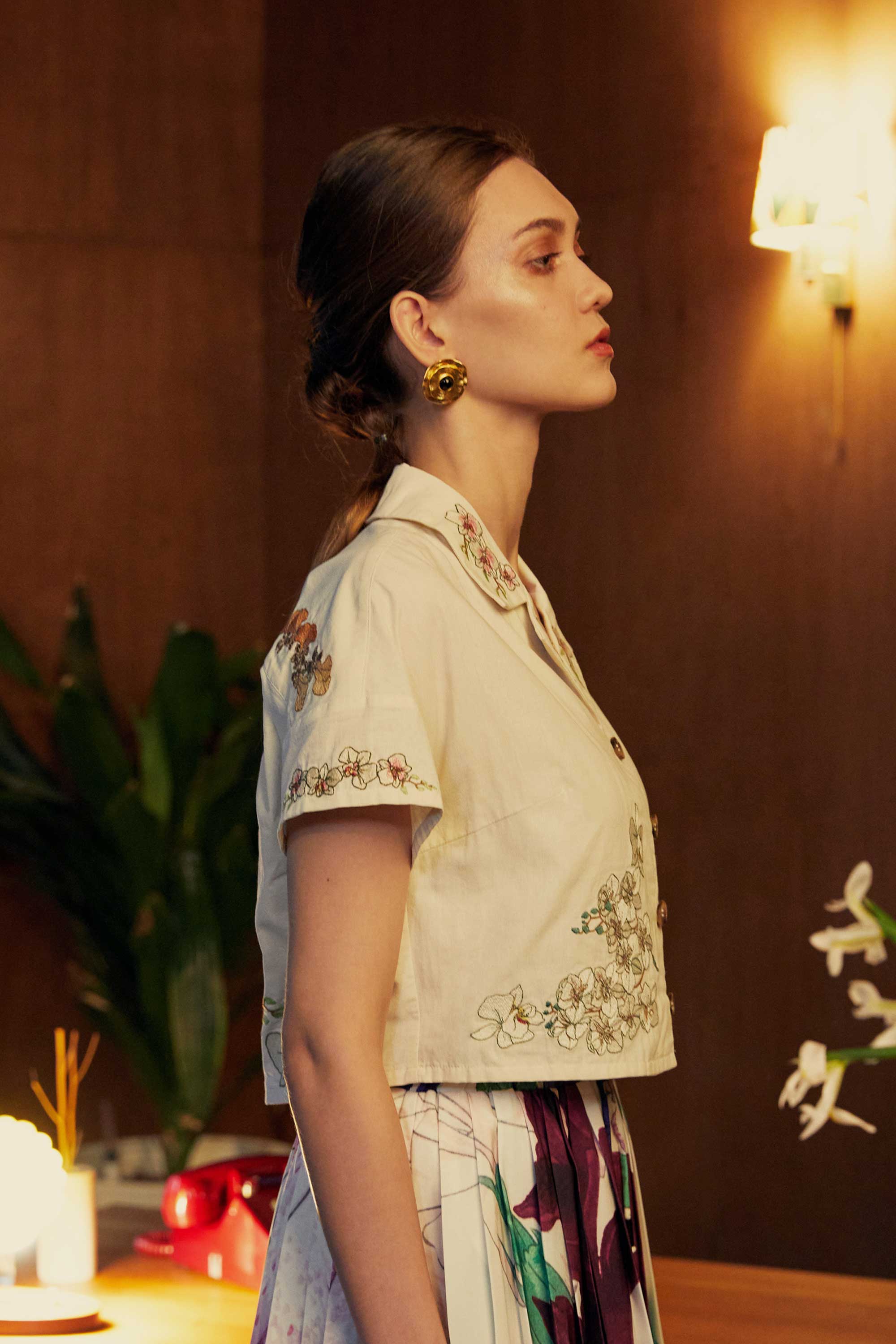 Cropped side profile of a model wearing a floral embroidered Cuban shirt and printed pleated skorts, styled against a wooden backdrop with plants and soft ambient lighting.