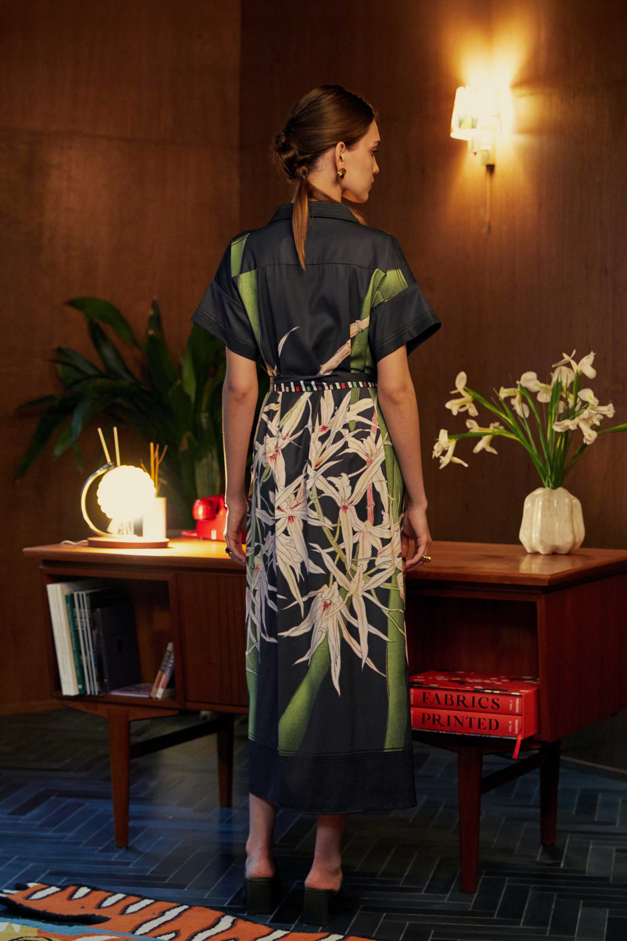 Back view of model in the BEHOLD Cuban Maxi Dress, Brassavola Cucullata orchid print in black and green, button-down maxi with belted waist and bold floral motif, styled indoors with wood-paneled backdrop and mid-century desk.