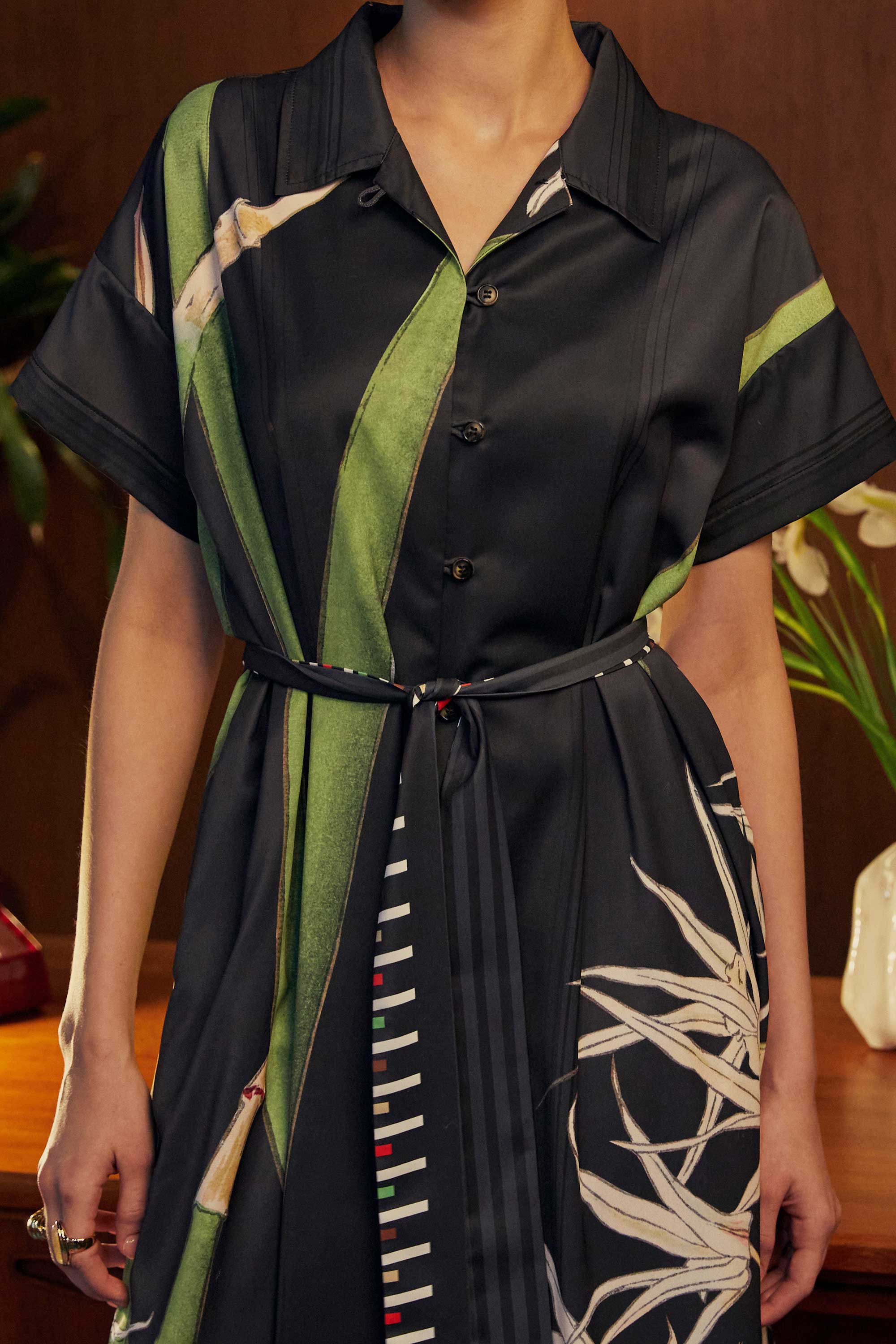 Close-up of the BEHOLD Cuban Maxi Dress in Brassavola Cucullata orchid print, black and green shirt-dress with button-down front, short sleeves, belted waist, and white orchid floral motifs.