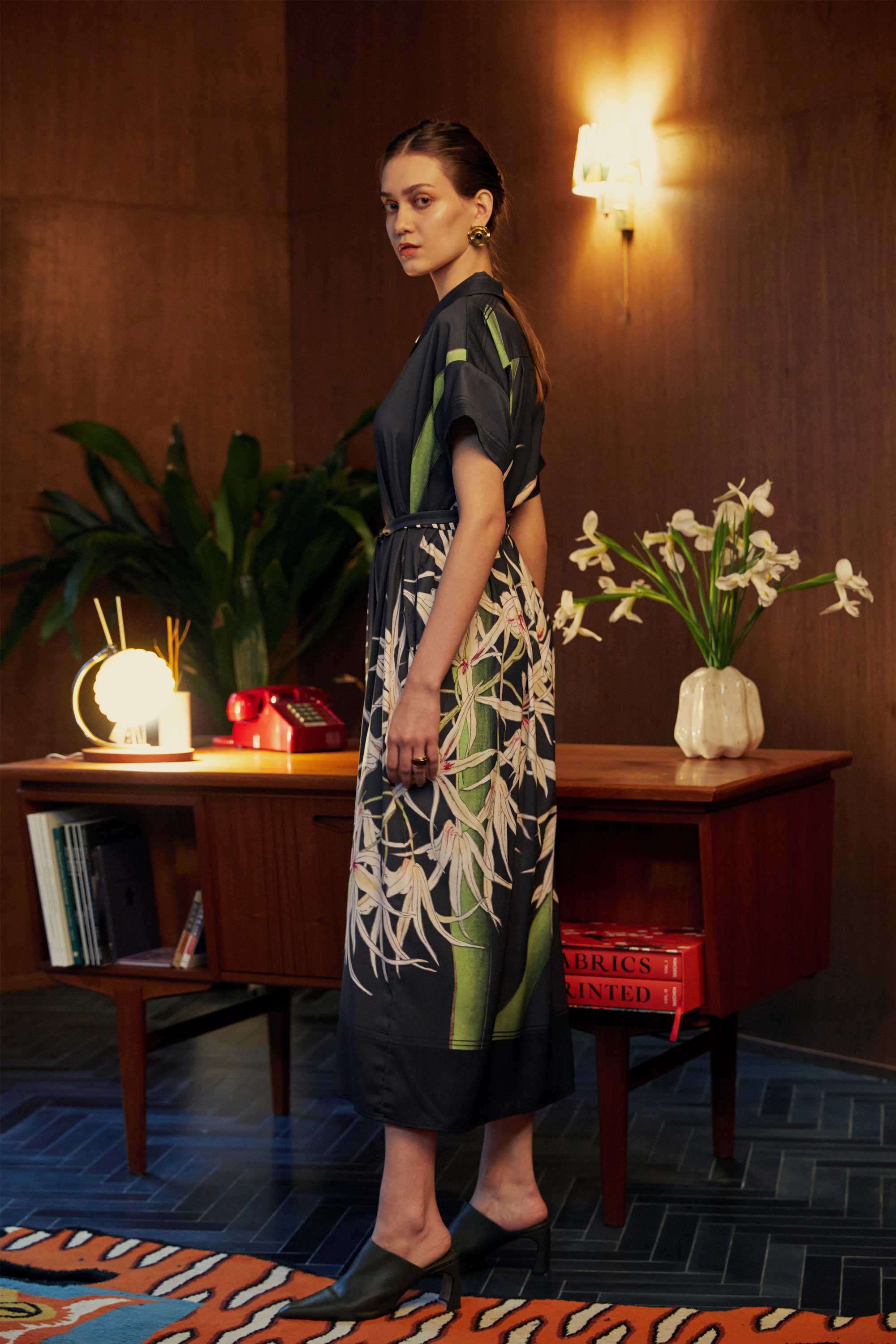 Side view of model in the BEHOLD Cuban Maxi Dress, Brassavola Cucullata orchid print in black and green, maxi shirt-dress with belted waist and floral motifs, photographed indoors with wood-panel background and desk.
