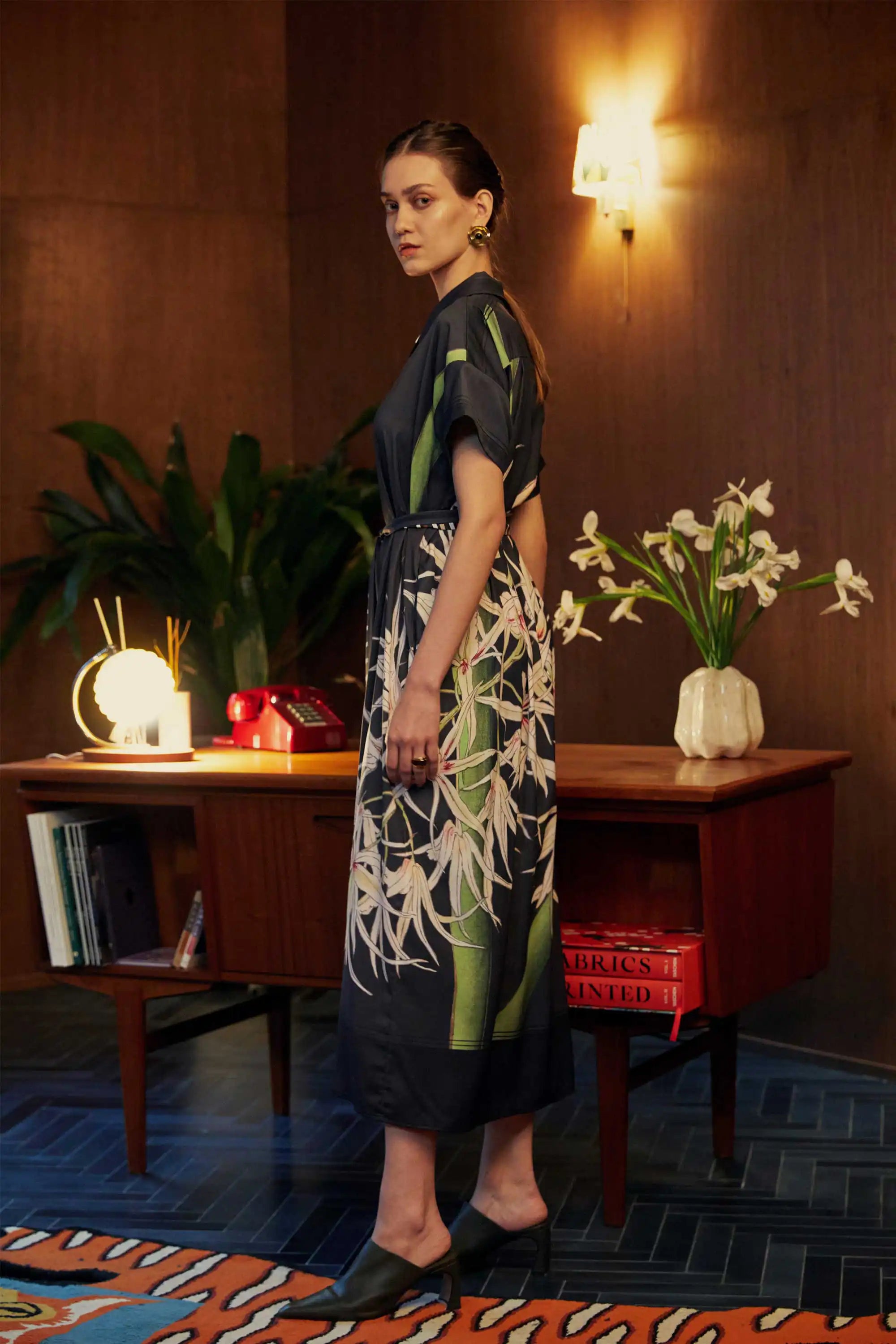 Side view of model in the BEHOLD Cuban Maxi Dress, Brassavola Cucullata orchid print in black and green, maxi shirt-dress with belted waist and floral motifs, photographed indoors with wood-panel background and desk.