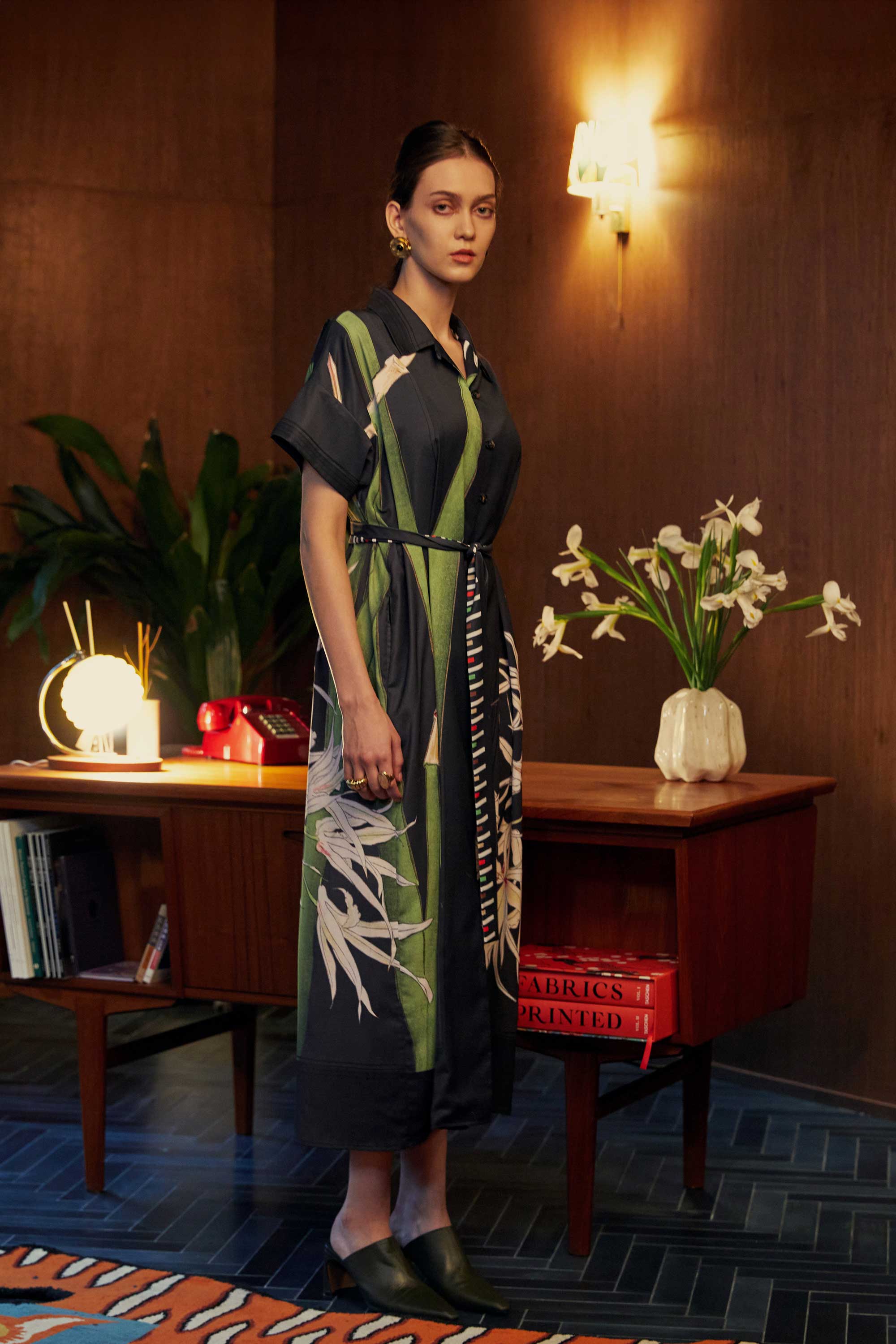 Side profile of model in the BEHOLD Cuban Maxi Dress, Brassavola Cucullata orchid print in black and green, button-down maxi with belted waist and floral detailing, styled indoors against wood-paneled walls with mid-century desk and decor.