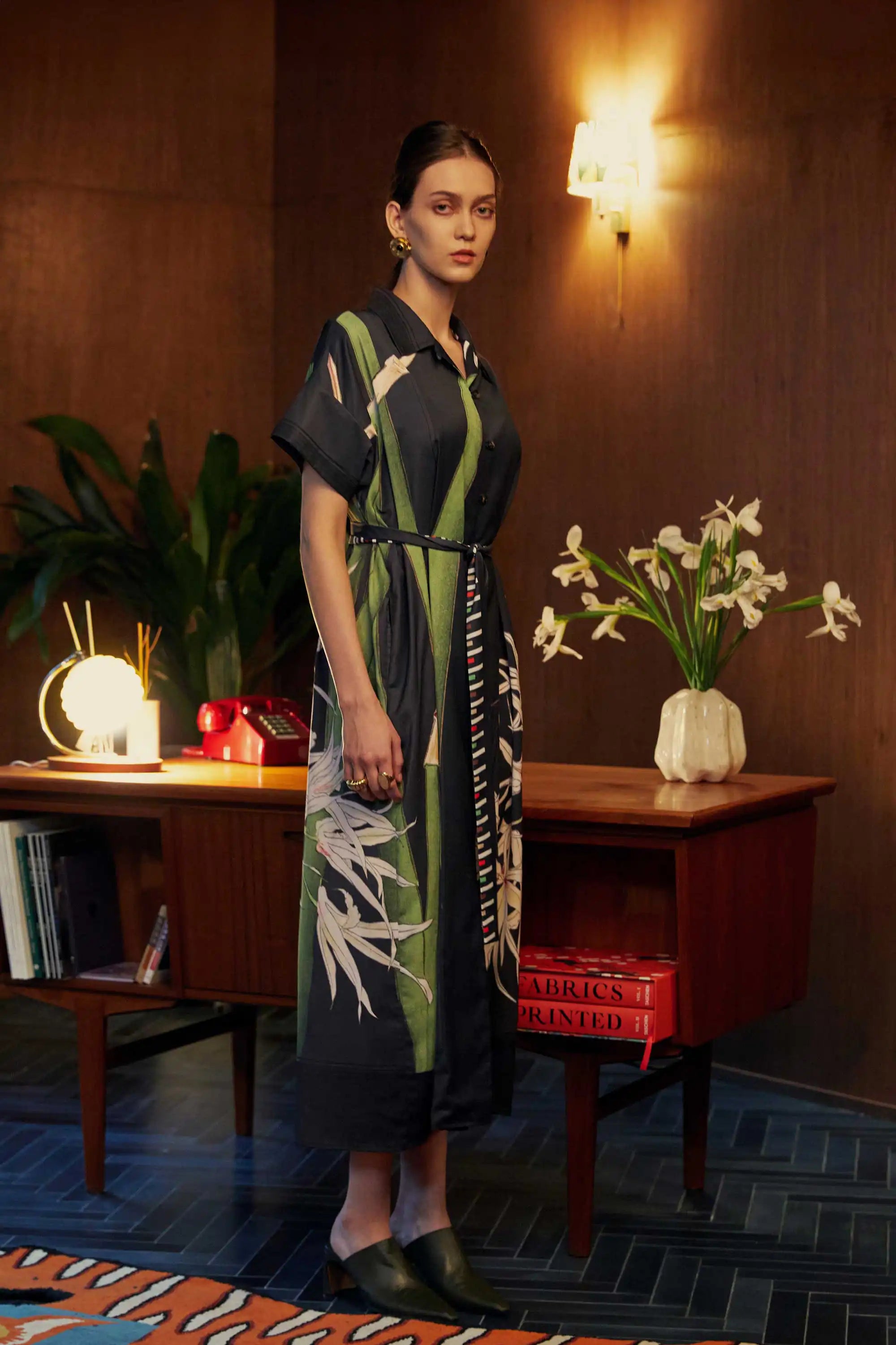 Side profile of model in the BEHOLD Cuban Maxi Dress, Brassavola Cucullata orchid print in black and green, button-down maxi with belted waist and floral detailing, styled indoors against wood-paneled walls with mid-century desk and decor.