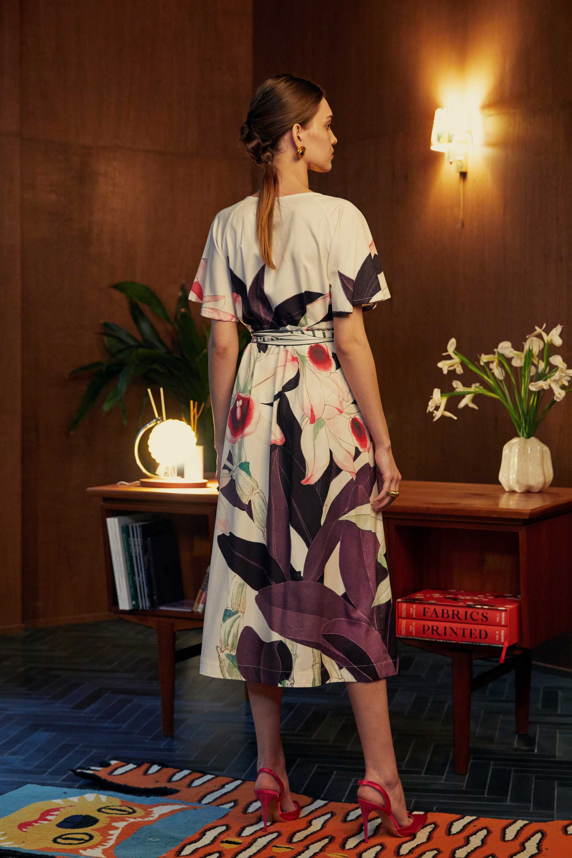 Back view of model in the BEHOLD Flutter Sleeves Box Pleats Cinch Midi Dress, Dendrobium orchid print on cream base with bold purple and pink floral motifs, flutter sleeves, and striped cinched waist, styled indoors with wood-paneled backdrop.