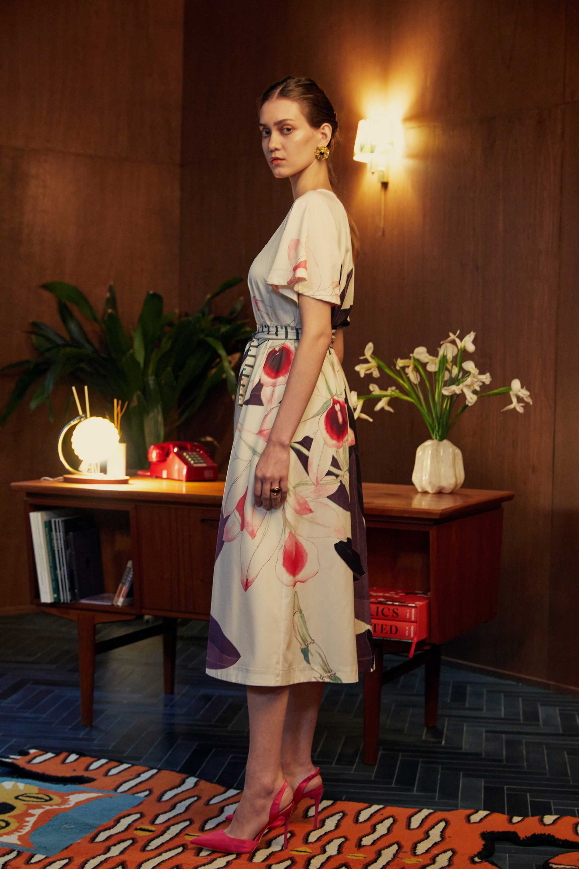 Side view of model in the BEHOLD Flutter Sleeves Box Pleats Cinch Midi Dress, Dendrobium orchid print on cream base, featuring flutter sleeves, striped cinched waist, and bold floral motifs, styled indoors with wood-paneled setting.