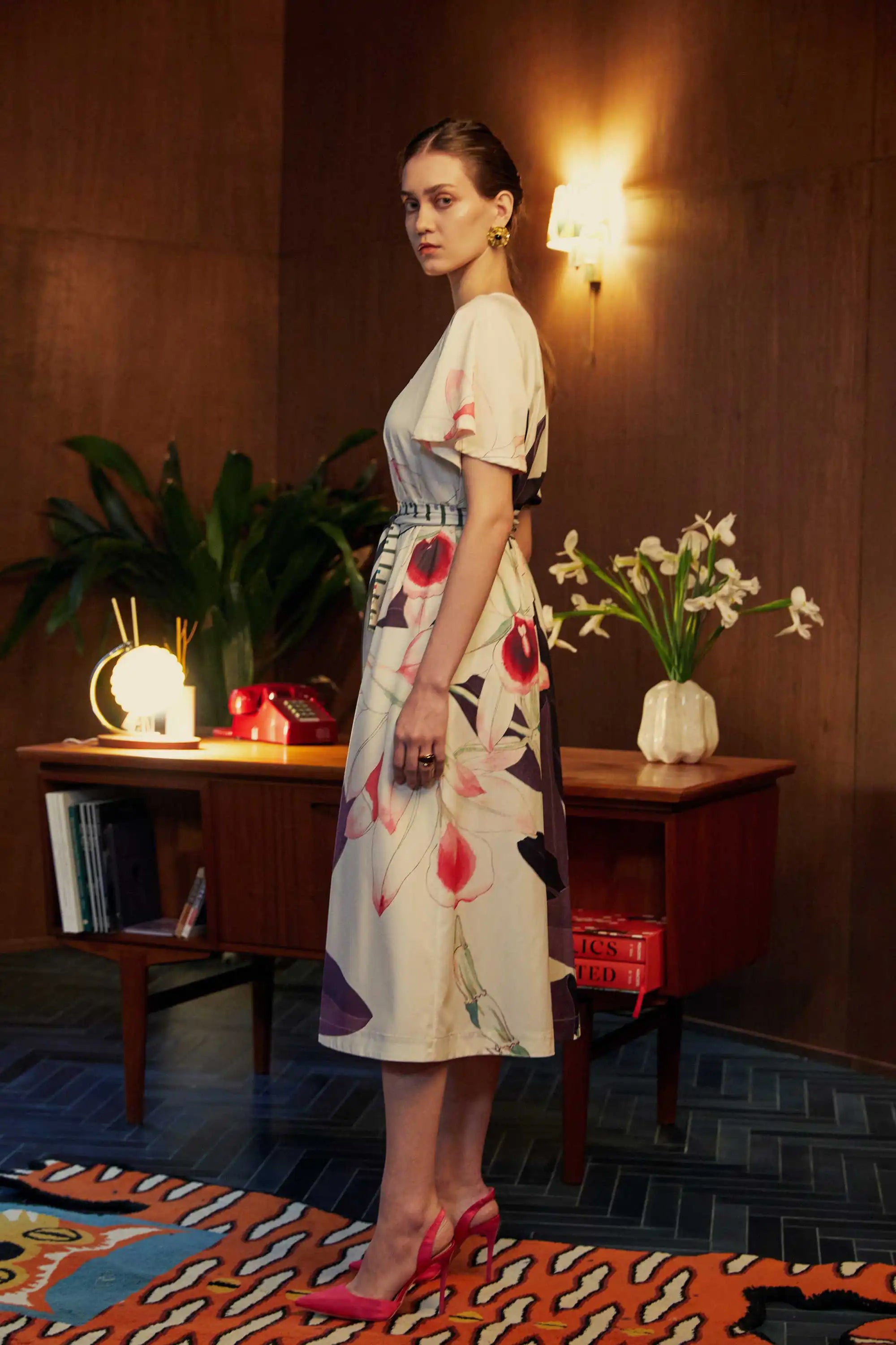 Side view of model in the BEHOLD Flutter Sleeves Box Pleats Cinch Midi Dress, Dendrobium orchid print on cream base, featuring flutter sleeves, striped cinched waist, and bold floral motifs, styled indoors with wood-paneled setting.