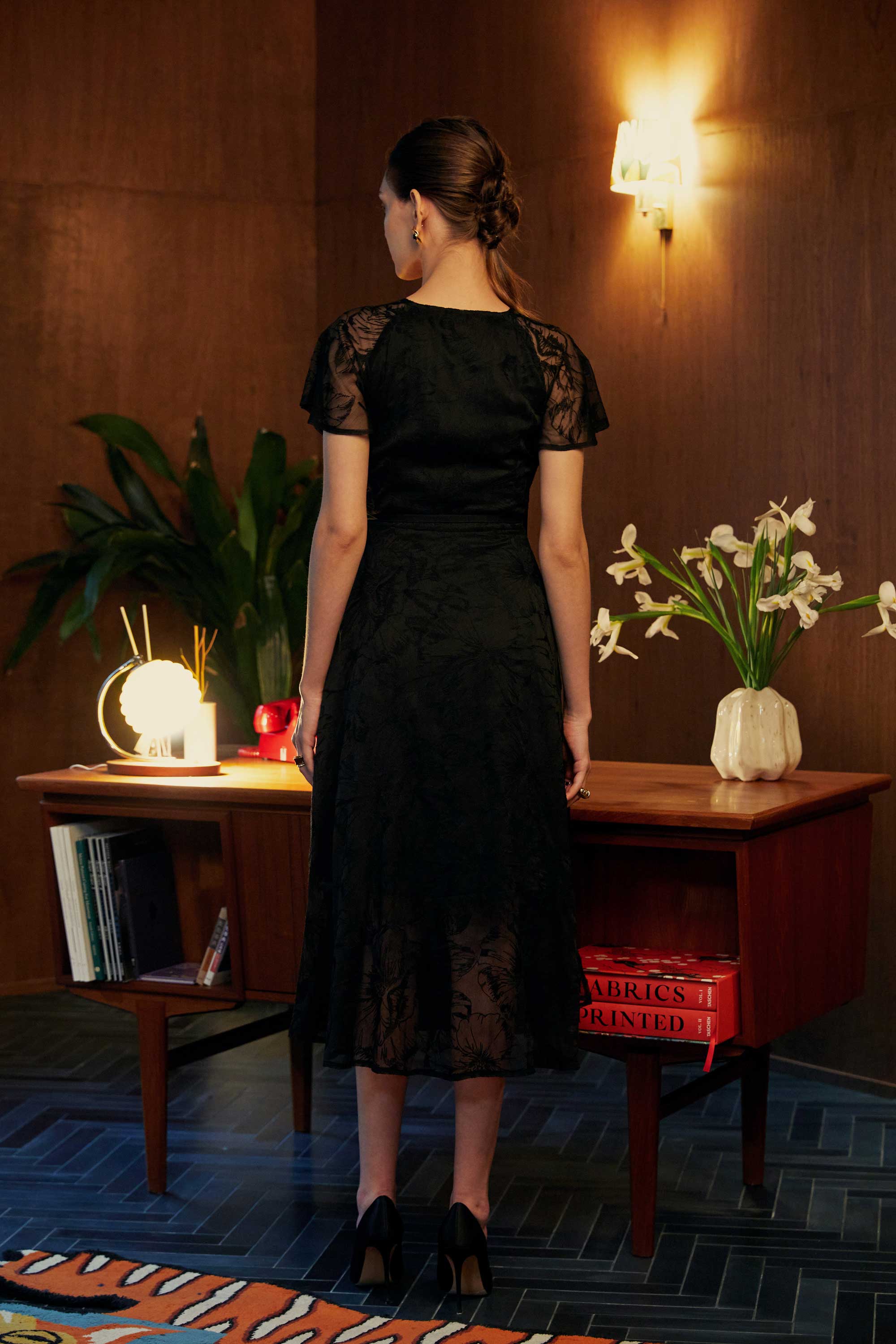 Back view of model in the BEHOLD Flutter Sleeves Wrap Midi Dress, black lace wrap midi with flutter sleeves and fitted waistline, styled indoors with wood-paneled backdrop and mid-century desk.