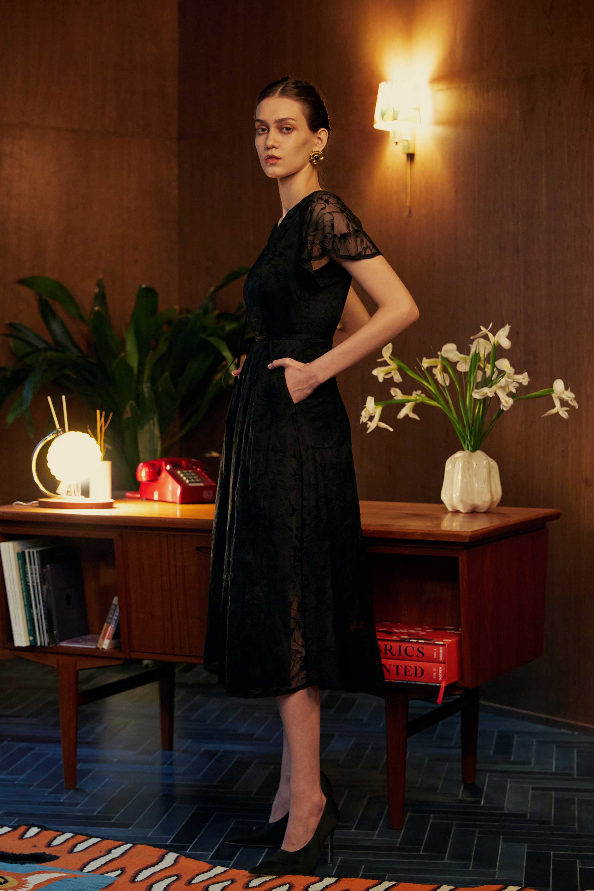 Side view of model in the BEHOLD Flutter Sleeves Wrap Midi Dress, black lace midi with flutter sleeves, V-neckline, and wrap silhouette, styled indoors with wood-paneled walls and mid-century desk.
