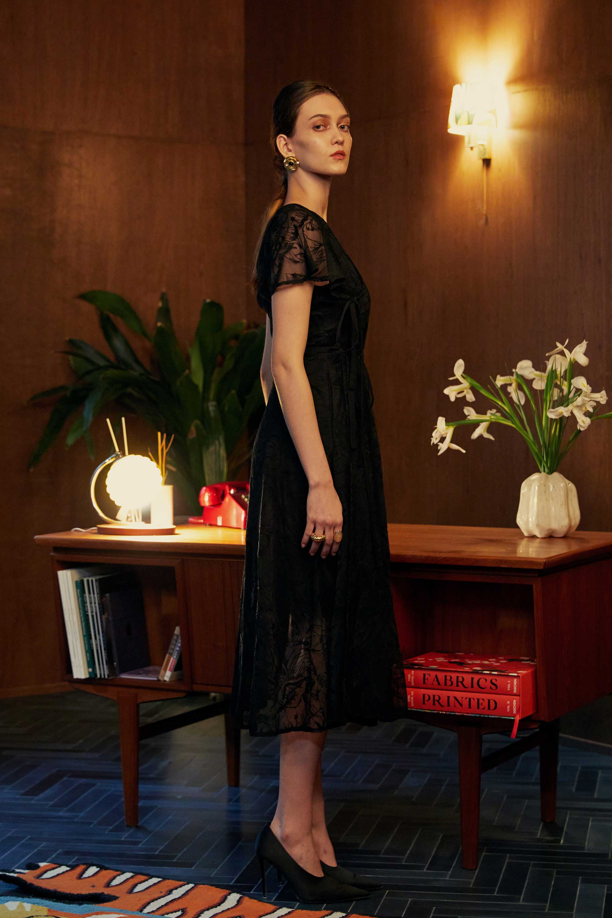 Side profile of model in the BEHOLD Flutter Sleeves Wrap Midi Dress, black lace wrap silhouette with flutter sleeves and V-neckline, styled indoors with wood-paneled background and decorative desk.