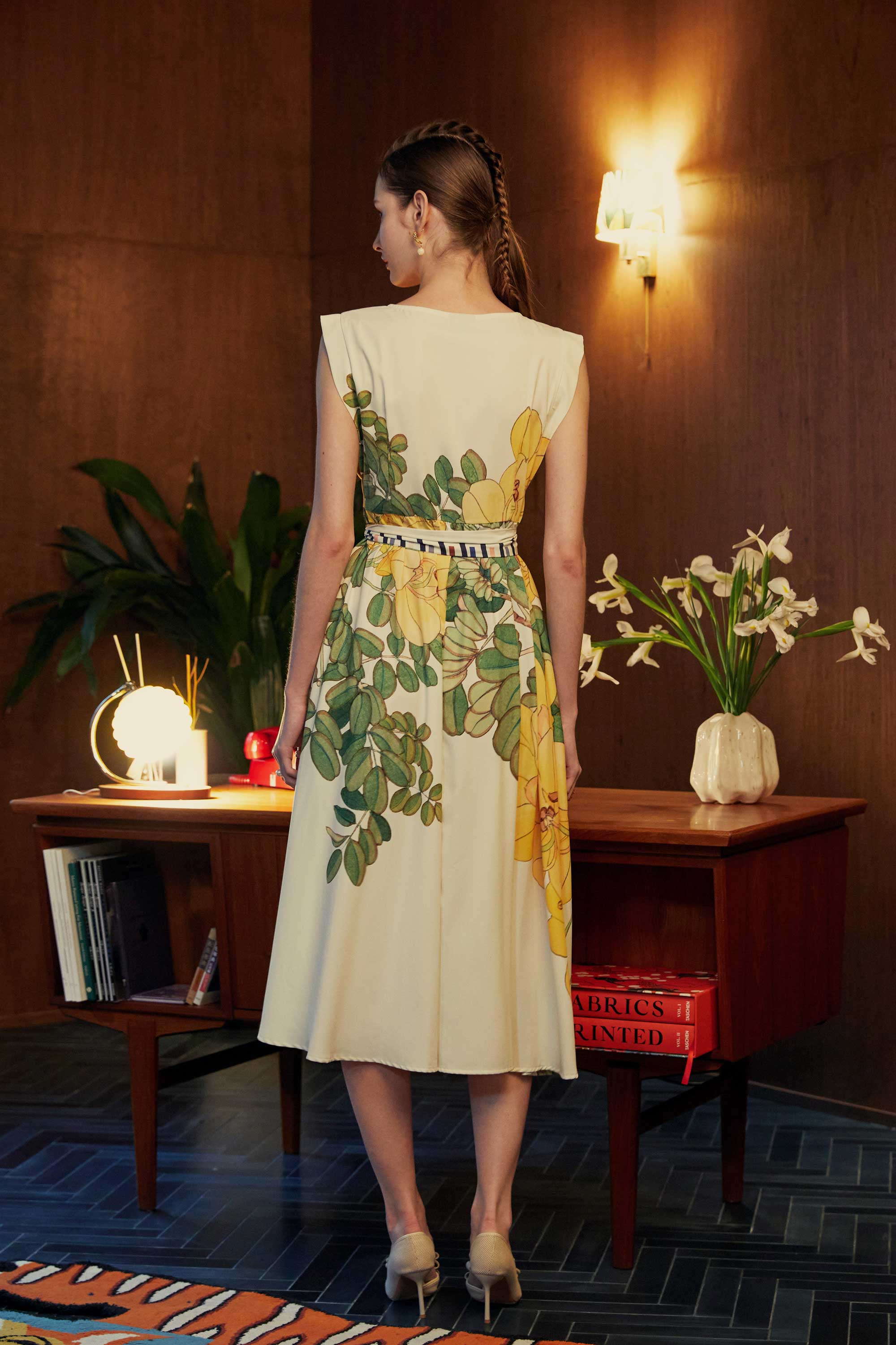 Back view of model in BEHOLD Origami Square Sleeve Dress featuring yellow allamanda florals, standing in a retro-inspired room with warm lighting and wooden furniture.