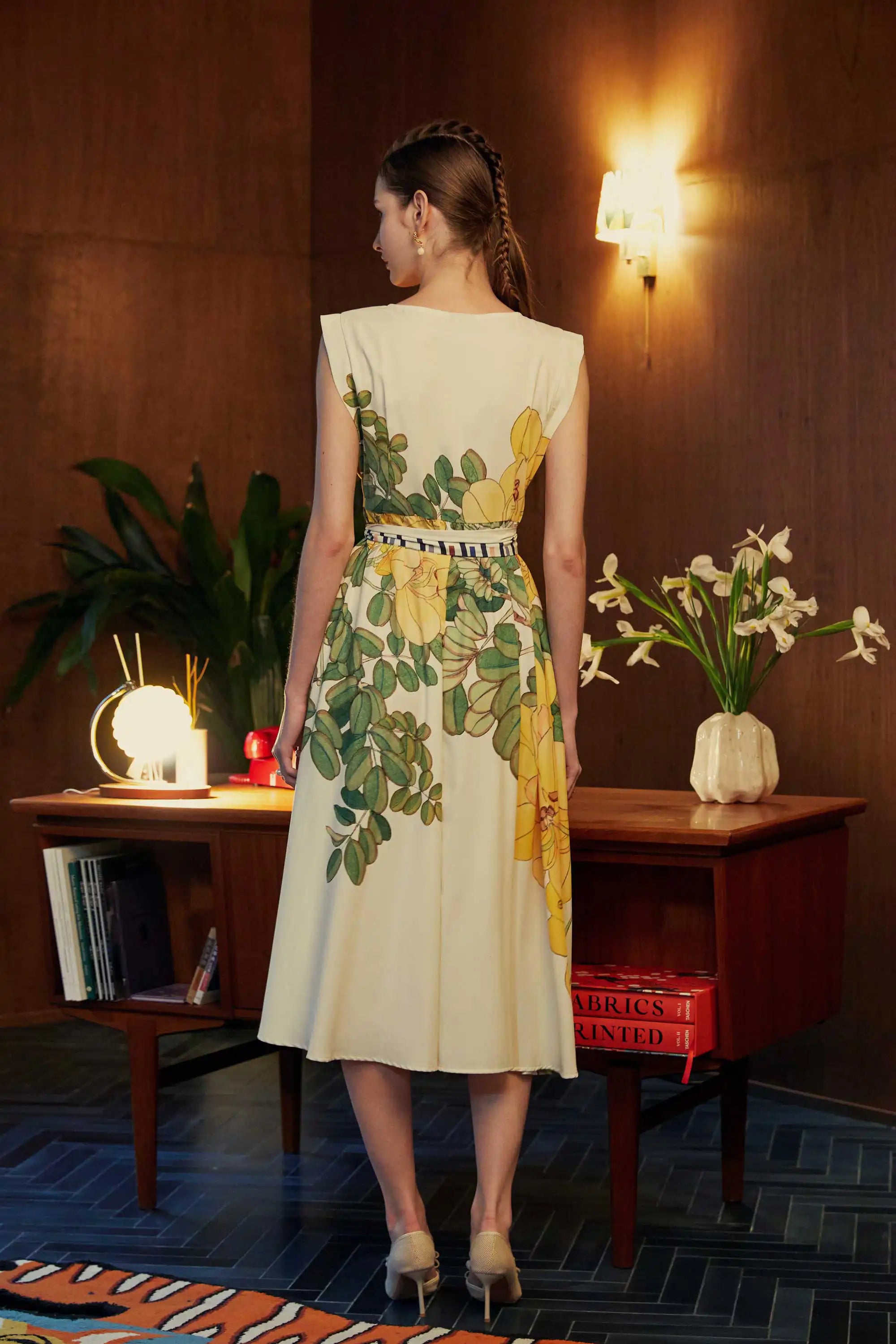 Back view of model in BEHOLD Origami Square Sleeve Dress featuring yellow allamanda florals, standing in a retro-inspired room with warm lighting and wooden furniture.