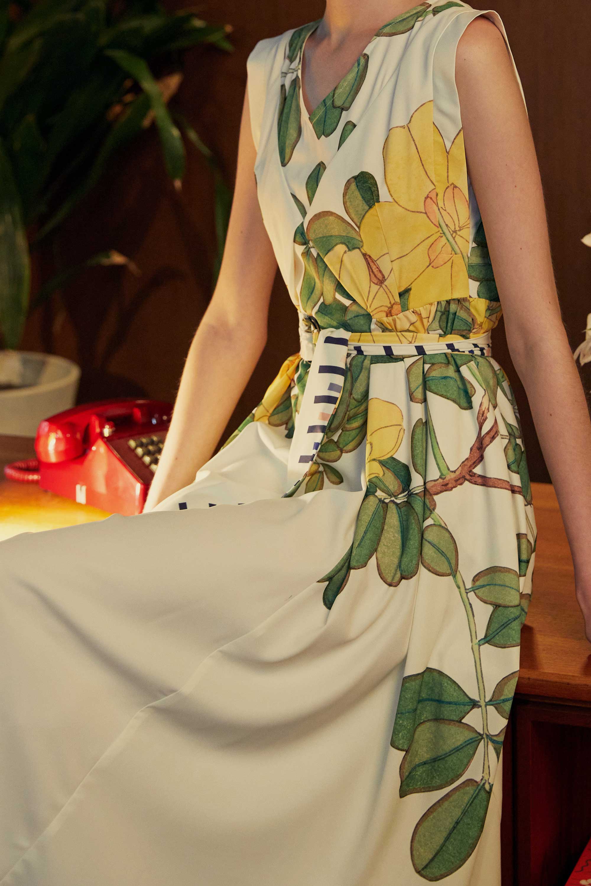 Side view of woman seated in BEHOLD Origami Square Sleeve Dress with Allamanda floral print, highlighting waist gathers, striped sash, and vibrant botanical motifs.
