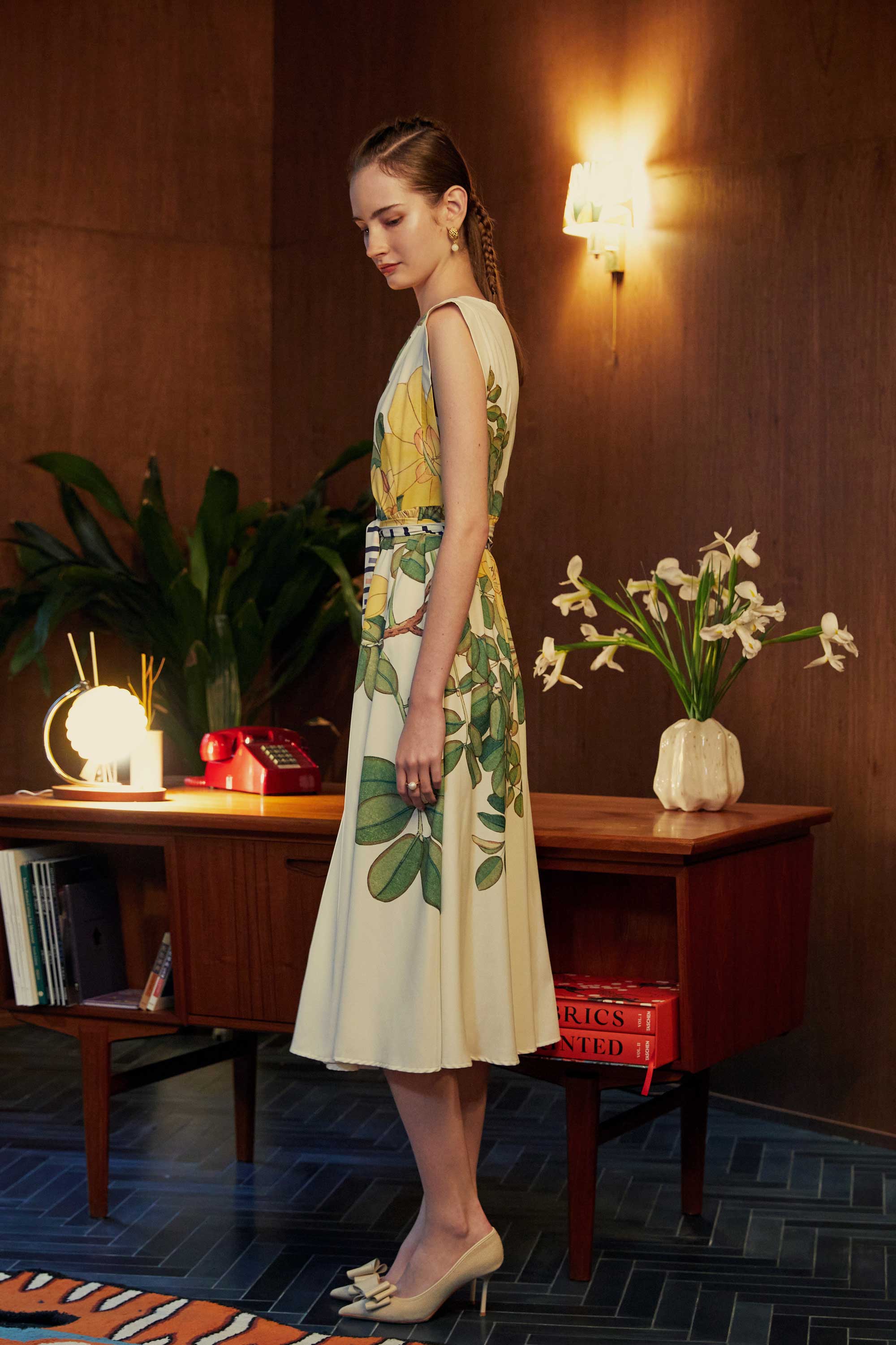 Side view of model in BEHOLD Origami Square Sleeve Dress featuring yellow allamanda floral print and striped belt, styled in a room with vintage decor and houseplants.