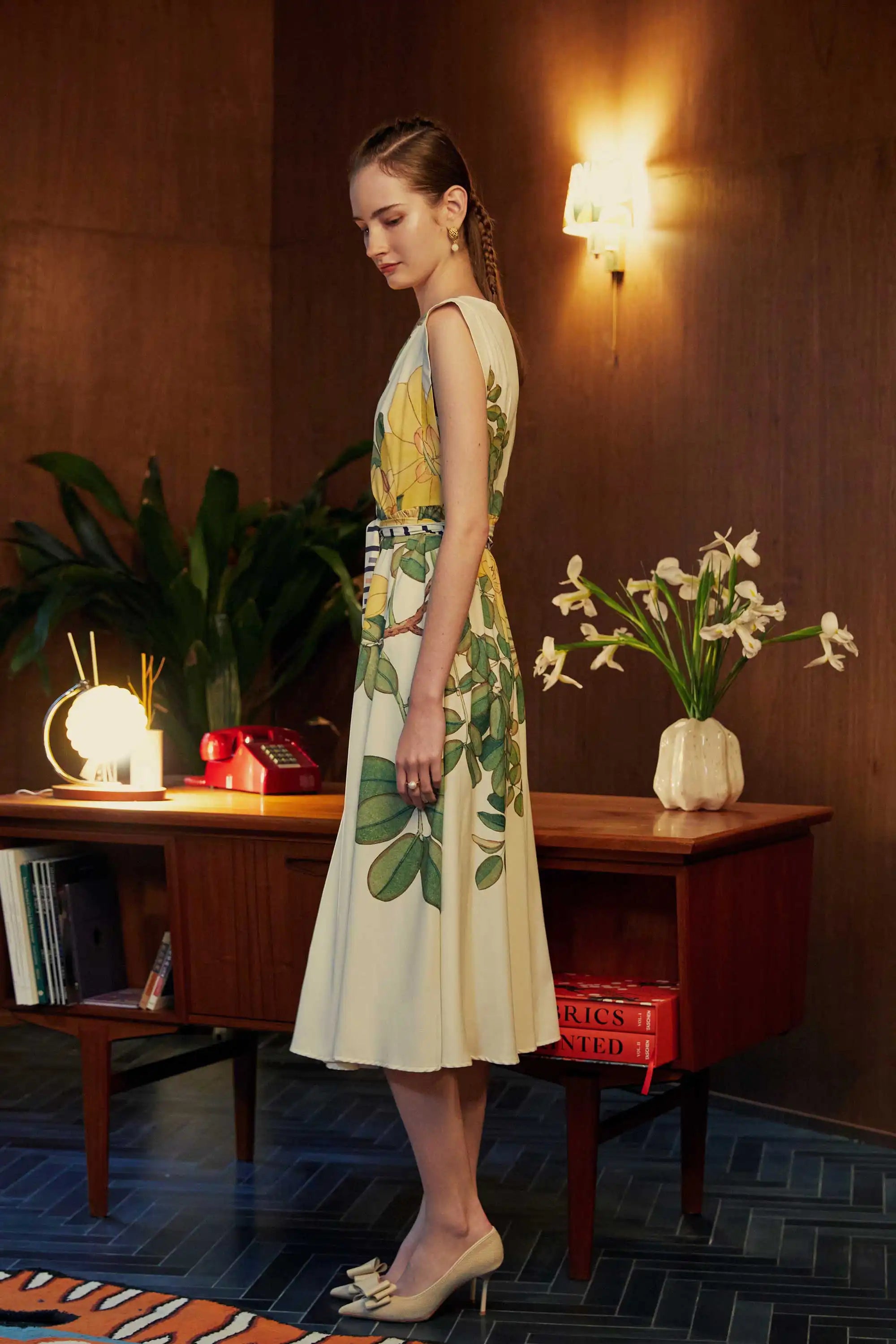 Side view of model in BEHOLD Origami Square Sleeve Dress featuring yellow allamanda floral print and striped belt, styled in a room with vintage decor and houseplants.