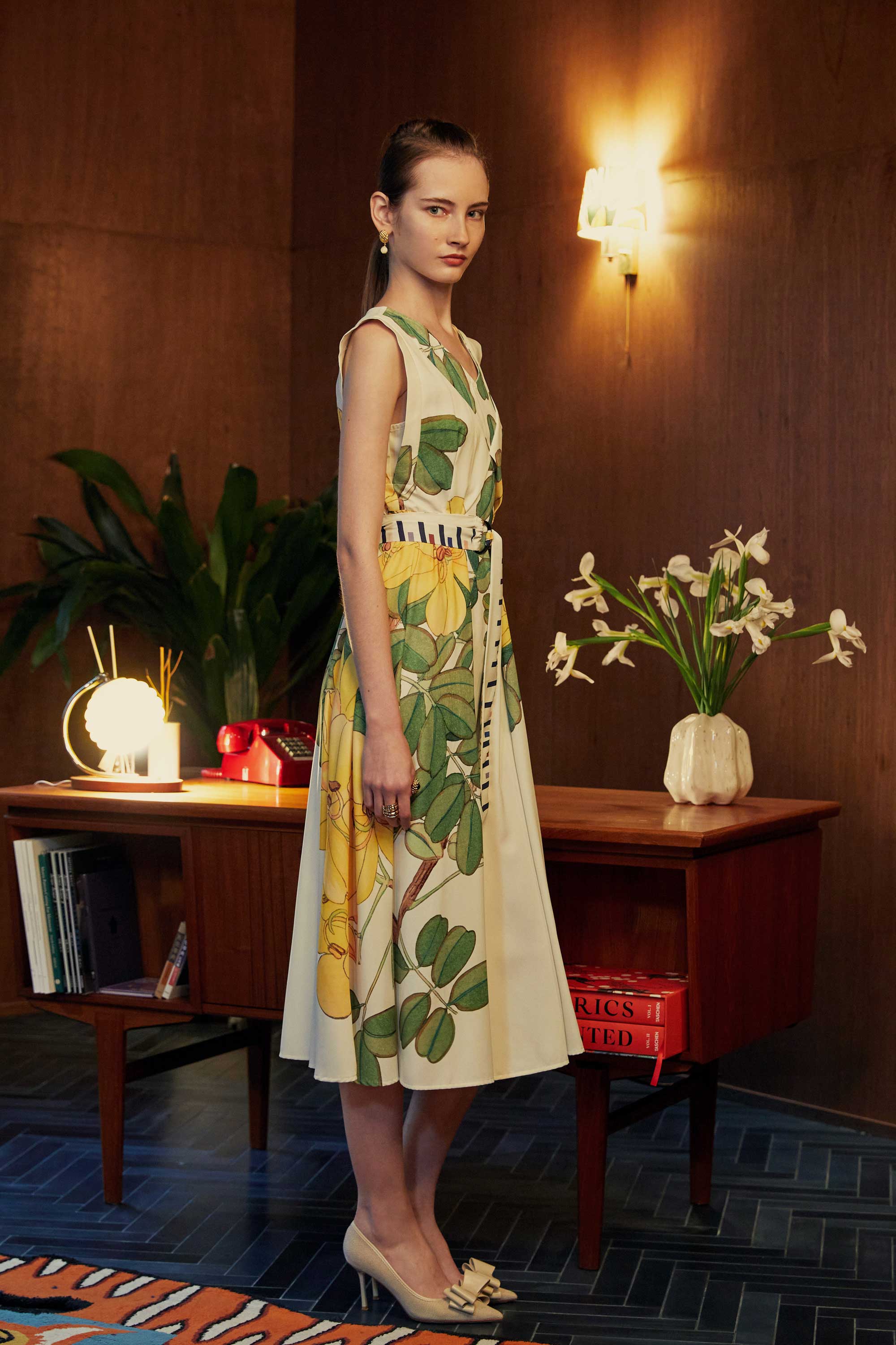 Model in BEHOLD Origami Square Sleeve Dress with vibrant yellow allamanda florals, viewed from the side, styled with neutral heels in a mid-century modern room.