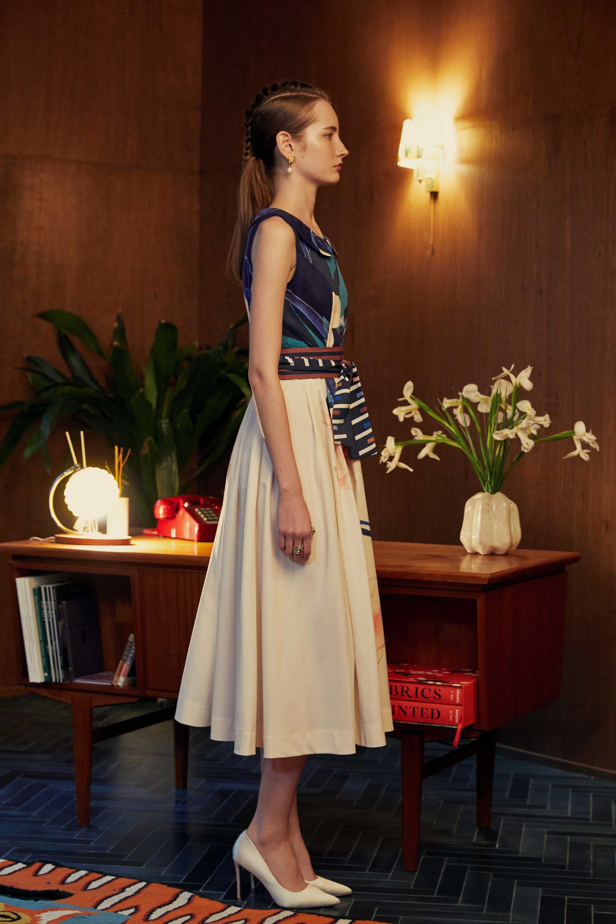 Side profile of model in the BEHOLD Peter Pan Fit-and-Flare Midi Dress in Dendrobium Chrysotoxum orchid print, featuring navy sleeveless bodice with Peter Pan collar, cream flared midi skirt with floral motifs, and striped waistband, styled indoors with wood-paneled background.