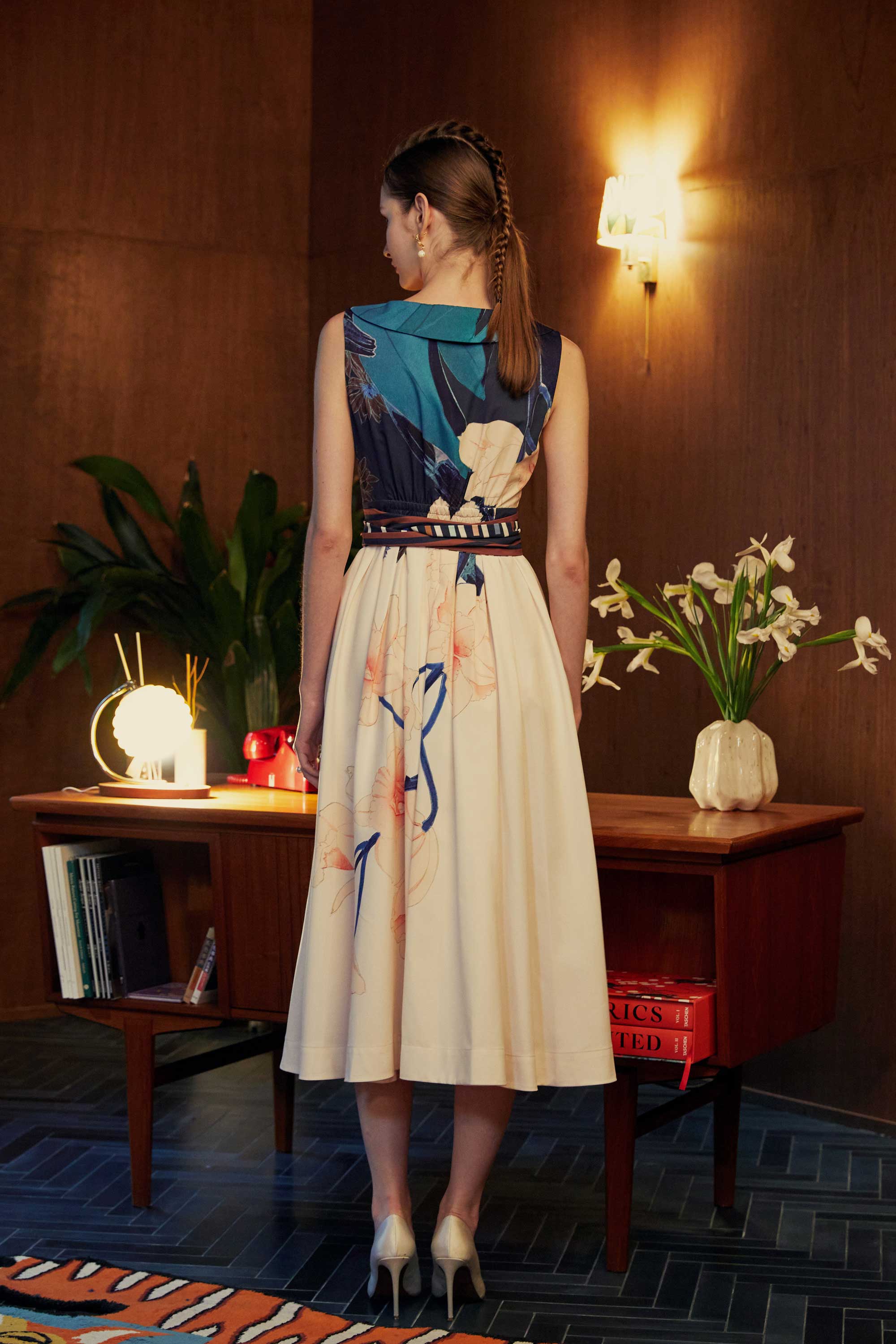 Back view of model in the BEHOLD Peter Pan Fit-and-Flare Midi Dress in Dendrobium Chrysotoxum orchid print, featuring navy sleeveless bodice, cream flared midi skirt with orchid floral motifs, and striped waistband, styled indoors with wood-paneled backdrop.