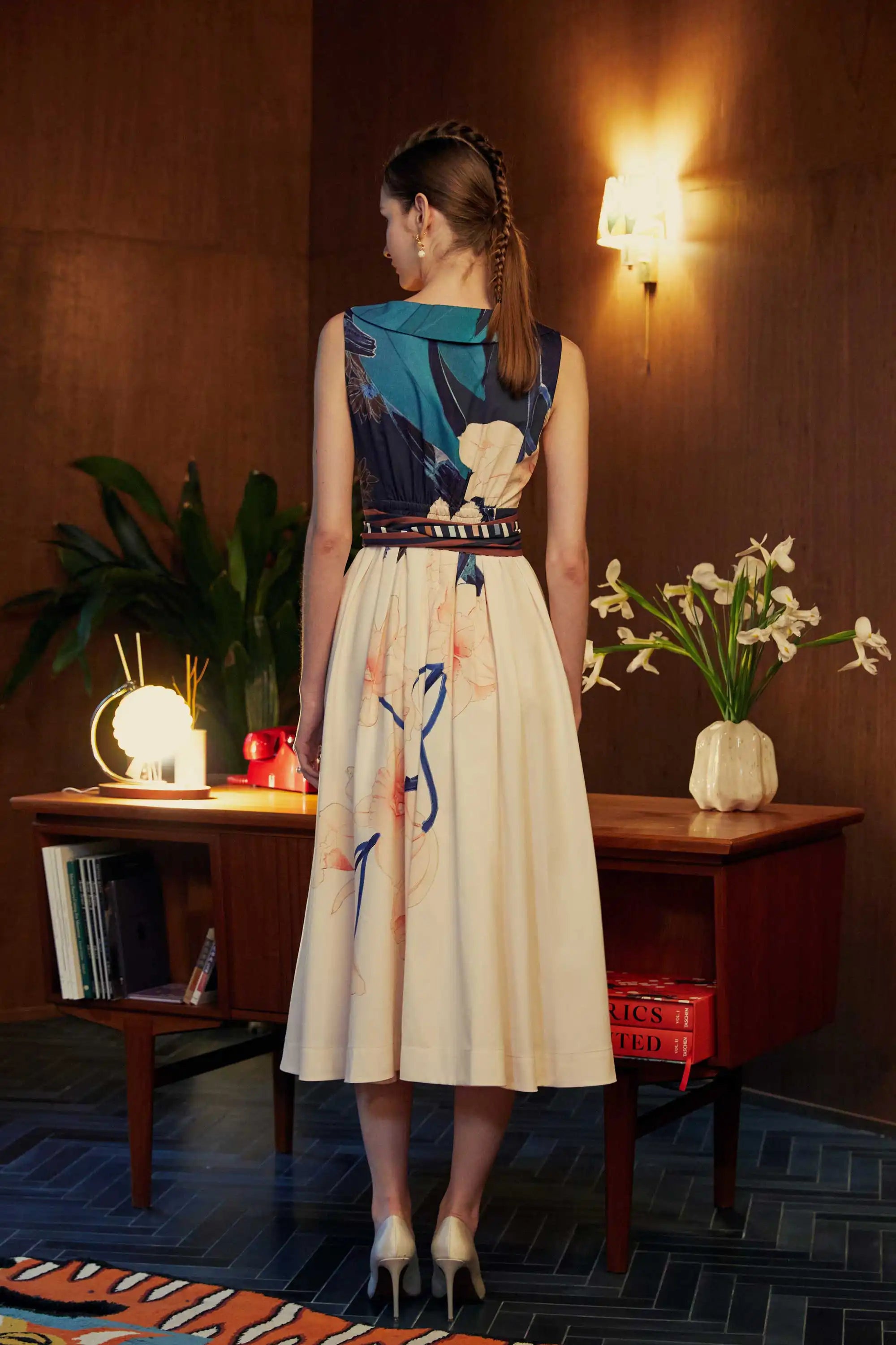 Back view of model in the BEHOLD Peter Pan Fit-and-Flare Midi Dress in Dendrobium Chrysotoxum orchid print, featuring navy sleeveless bodice, cream flared midi skirt with orchid floral motifs, and striped waistband, styled indoors with wood-paneled backdrop.