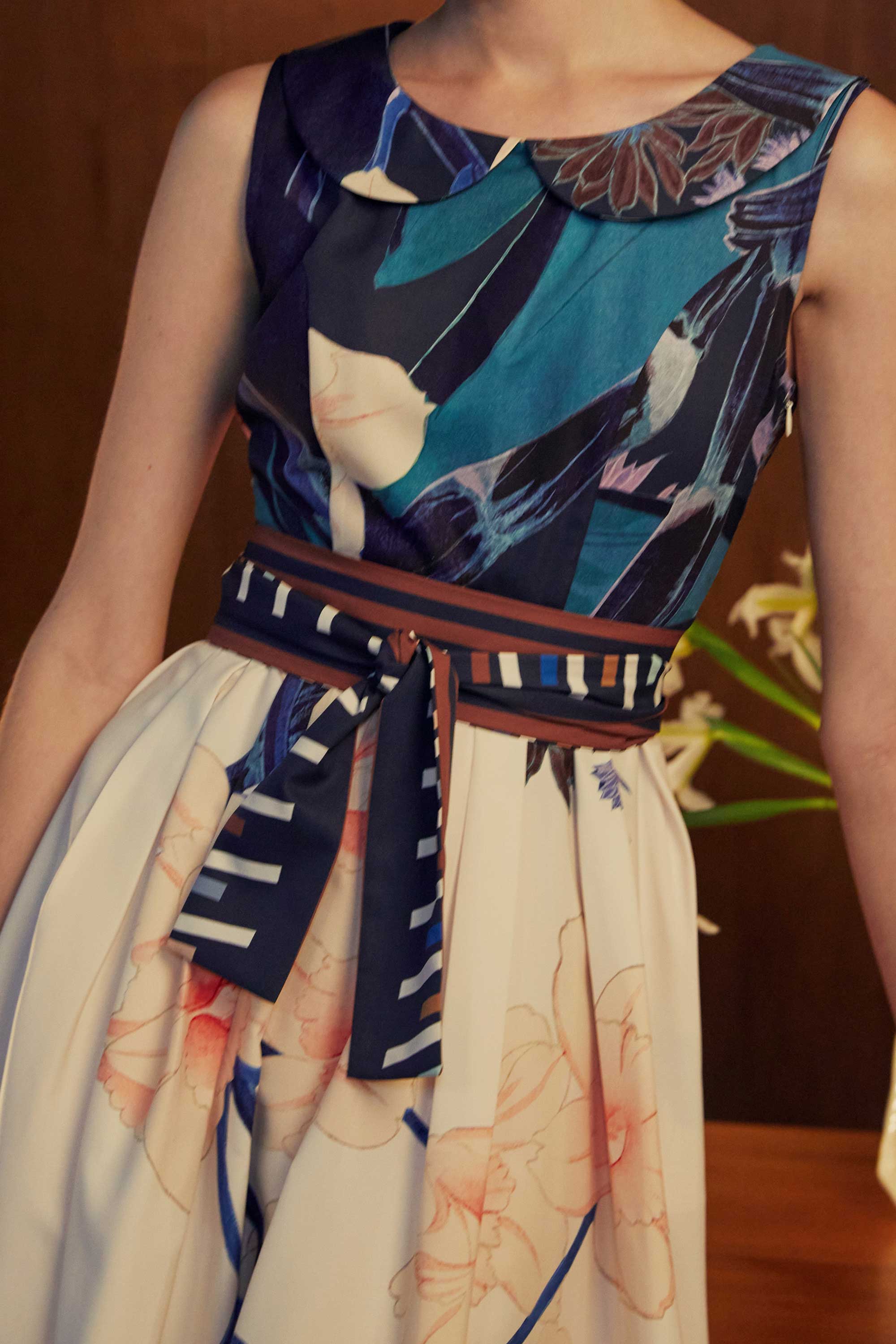 Close-up of the BEHOLD Peter Pan Fit-and-Flare Midi Dress in Dendrobium Chrysotoxum orchid print, highlighting navy sleeveless bodice with Peter Pan collar, cream skirt with orchid motifs, and striped waistband tie detail.