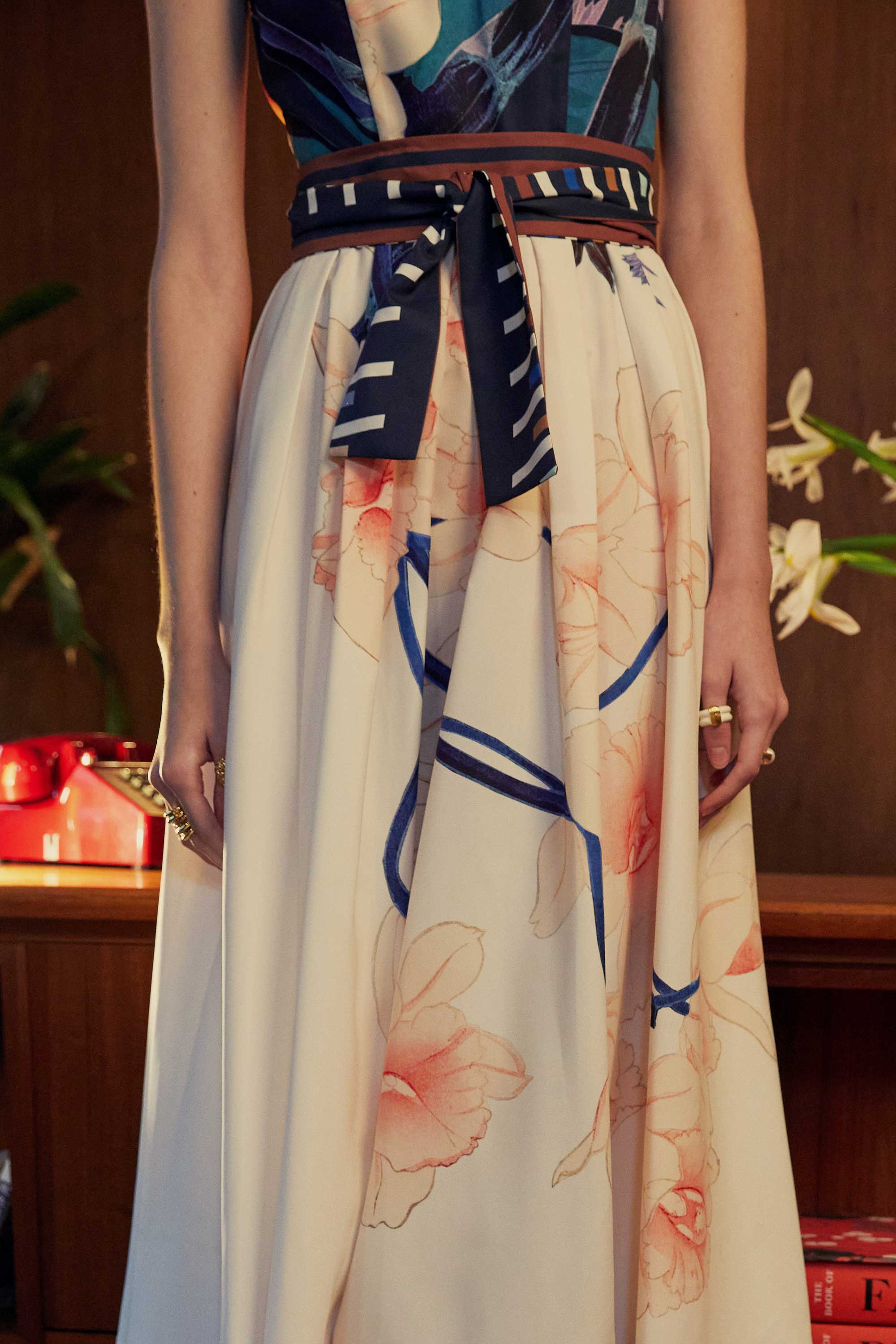 Detail view of the BEHOLD Peter Pan Fit-and-Flare Midi Dress in Dendrobium Chrysotoxum orchid print, showing striped waistband tie, cream flared skirt with soft orchid floral motifs, and navy sleeveless bodice.
